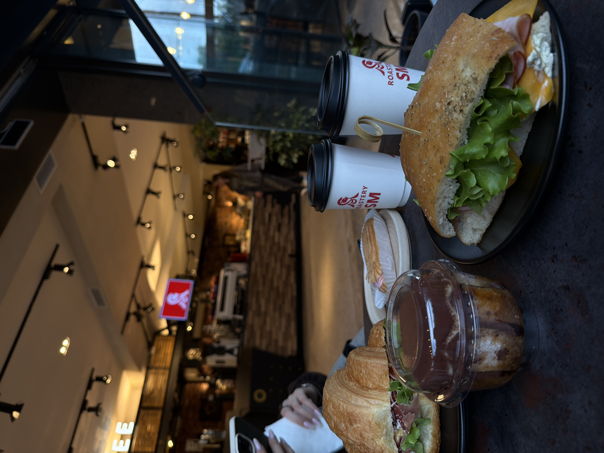 On a dark, possibly metallic table in an unknown city, a partially obscured view of a person with manicured fingernails using a smartphone, likely at a cafe or restaurant. On the table, there are two white cups with a red logo and black lids, positioned near a sandwich. The sandwich appears to be made with lettuce, ham, cheese, and mayonnaise. There is also a croissant-like pastry with green lettuce, ham, and what appears to be chocolate in a plastic container. In the background, the interior of the cafe includes a sign with a logo, dark wooden shelves, and ceiling lights. The view through the window shows buildings in the distance.