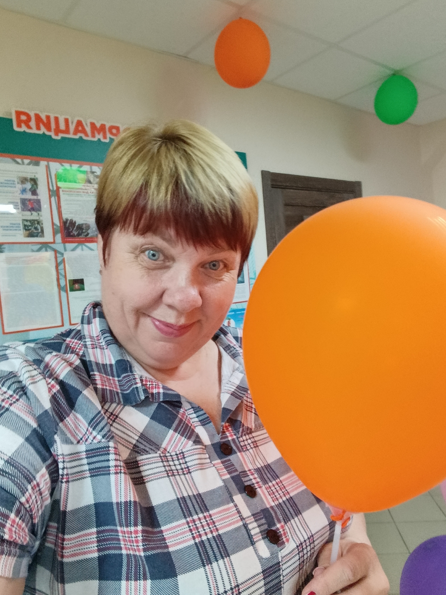 In the city of Svietlahorsk, Belarus, a middle-aged woman with short blonde hair and blue eyes is captured in a selfie. She is wearing a plaid shirt in shades of blue, white, red, and gray. To her right, a large orange balloon dominates the foreground, held by her right hand. In the background, an orange balloon is visible on the ceiling to the left, and a green balloon is on the ceiling to the right. A bulletin board with several papers and images is mounted on the wall behind the woman, with the partial word "RNHAMO" visible on a sign above it. A wooden door is also present in the background. The overall setting appears to be an indoor event or celebration.