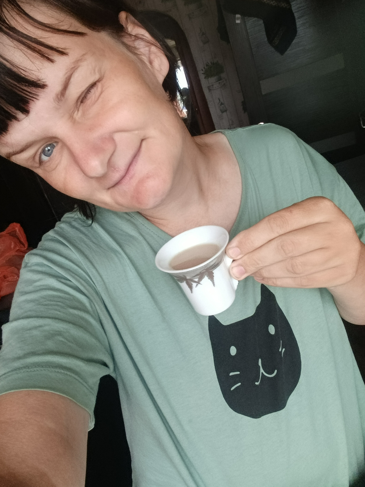 A woman winks at the camera while holding a small cup of coffee. She wears a light green t-shirt with a black cat design. The environment appears to be indoors, possibly a home, with a door and wallpaper visible in the background. The location is Фоки, Russia.