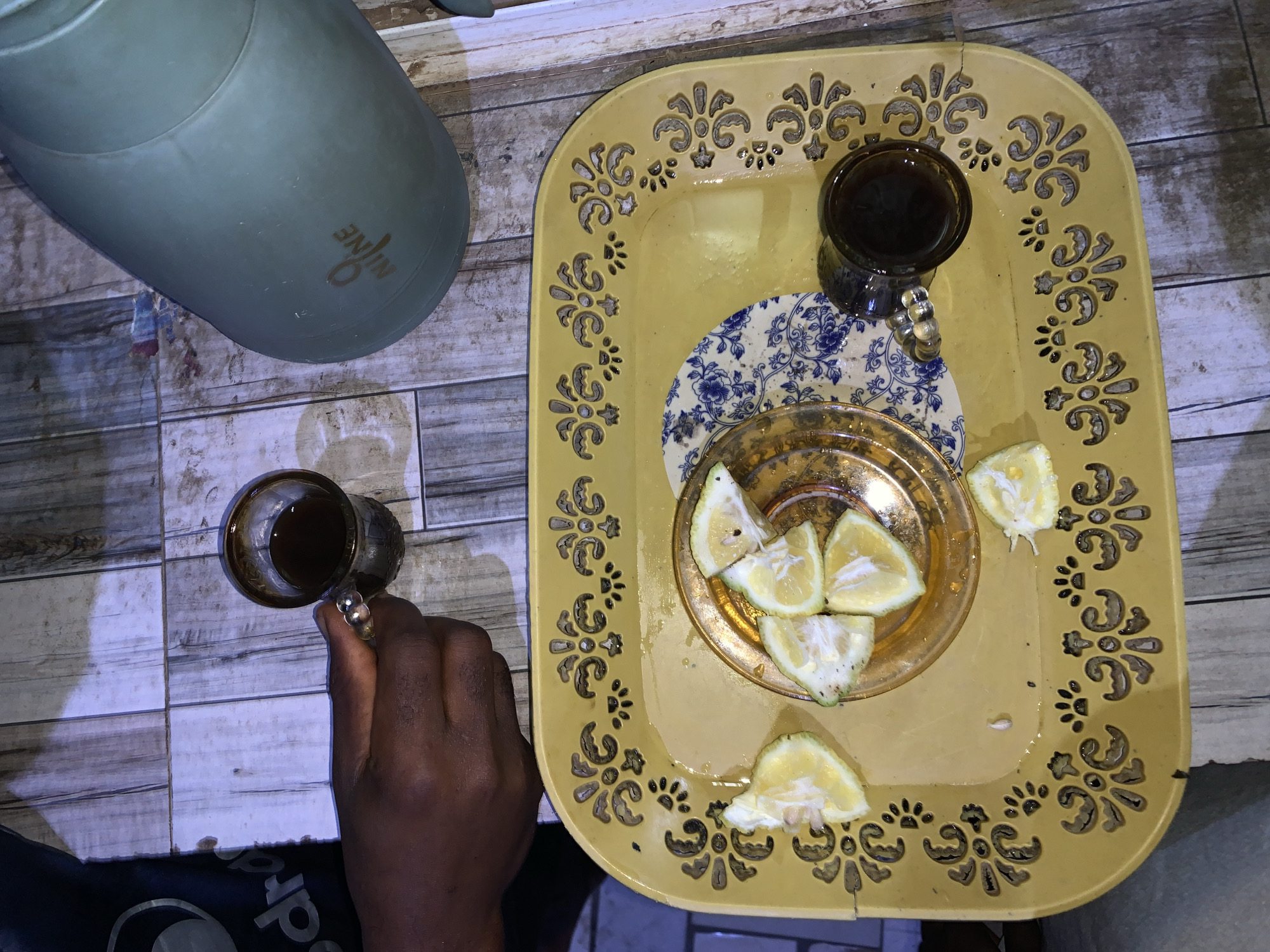 A top-down view captures a scene featuring a light green thermos next to a yellow tray. The tray holds a blue and white patterned saucer, a small glass of dark liquid (likely tea or coffee), and a gold plate with several slices of lemon. A hand is holding a similar glass of dark liquid. The location is inferred to be New Stadium, Nigeria. The environment appears to be an indoor space with a tiled surface.