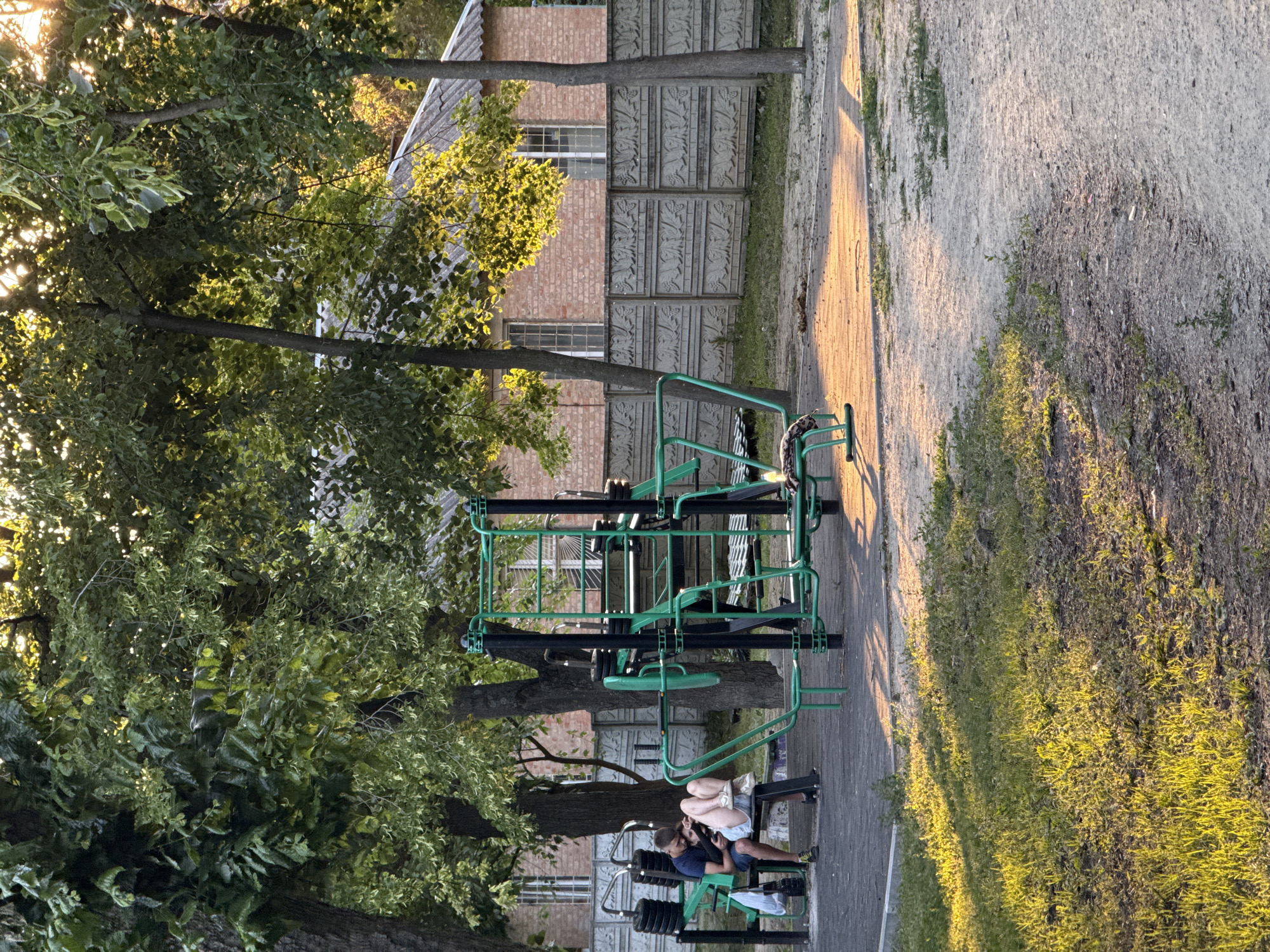 This is a medium shot of an outdoor exercise area with a young couple sitting on a bench. The man is wearing a dark blue t-shirt and shorts, and he is looking at a phone. The woman is wearing a light-colored top and denim shorts, and she is leaning her head on his shoulder, looking at the phone as well. They are sitting on a black bench positioned in front of weight stacks. To their right is a large, green and black outdoor weight training apparatus. Behind the bench and apparatus is a grey concrete fence with a decorative pattern. Beyond the fence, a brick building with a grey, corrugated roof and two windows is visible. The area is surrounded by numerous trees with green foliage, and the sunlight casts long shadows, indicating it is likely late afternoon or early morning. The ground in the foreground is a mixture of paved surfaces and dirt with grass.