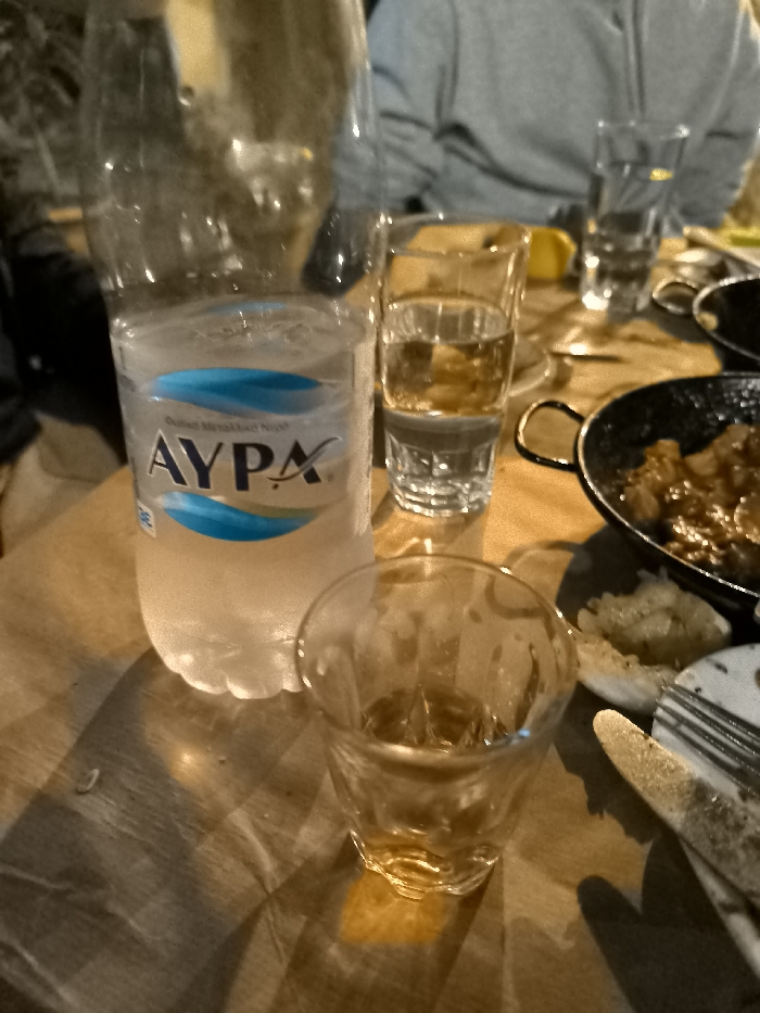 The image depicts a casual dining scene at an indoor restaurant or taverna, likely in Nea Erithrea, Greece, during the evening given the warm, artificial lighting. In the foreground, a plastic water bottle, approximately two-thirds full of ice-cold water, is prominent. The brand "AYPA" is clearly visible on its label in blue lettering, alongside other smaller, unreadable text.
Next to the water bottle, a small, clear glass holds a modest amount of an amber-colored liquid, possibly a spirit like ouzo or tsipouro, common in Greek tavernas. Further back, a taller glass also contains a clear beverage. To the right, a black frying pan with handles holds a portion of cooked food, appearing to be a hearty stew with pieces of meat and onions, indicative of traditional Greek cuisine. Adjacent to the pan, some bread or another food item rests on a plate, accompanied by a fork and a knife.
The table surface is a light-colored wood or a wood-patterned covering. In the blurred background, partial figures of people are visible, including one wearing a grey top, suggesting companions sharing the meal. The atmosphere is relaxed and social, typical of a casual dining experience. The dim, warm lighting creates a comfortable ambiance and casts soft shadows across the table.