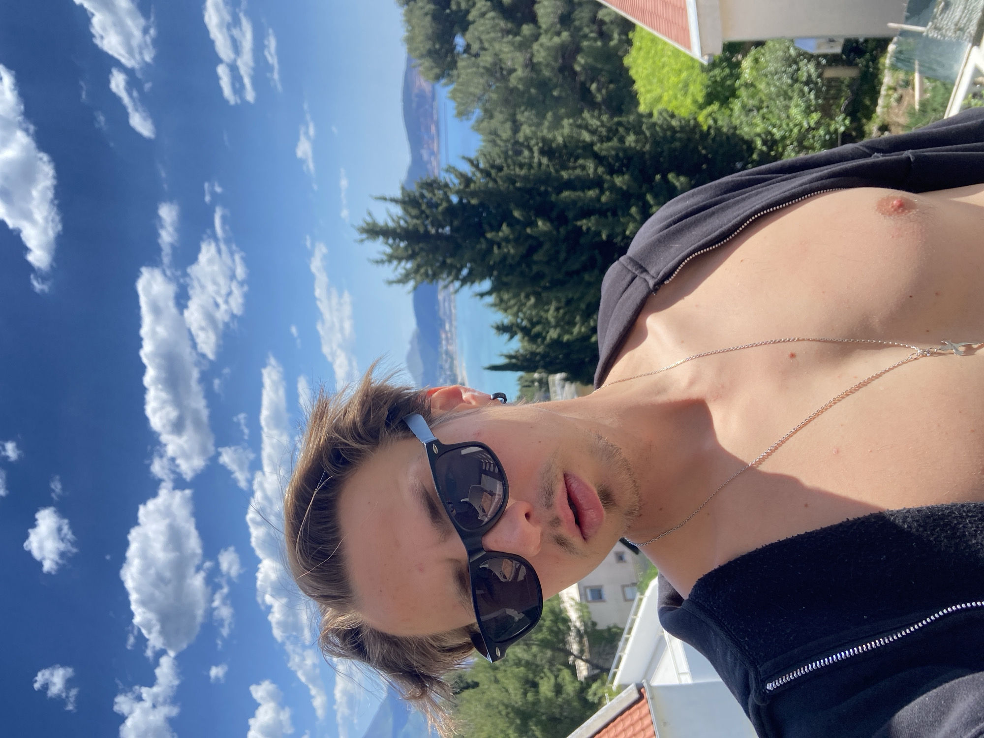 A man wearing sunglasses and a partially unzipped black jacket is outdoors in Šušanj, Montenegro, with a view of the sea and surrounding landscape. The sky is blue with scattered clouds. The background includes trees and buildings with red tile roofs. The man has fair skin, light brown hair, and a thin silver chain necklace. His jacket is partially open, exposing his chest.