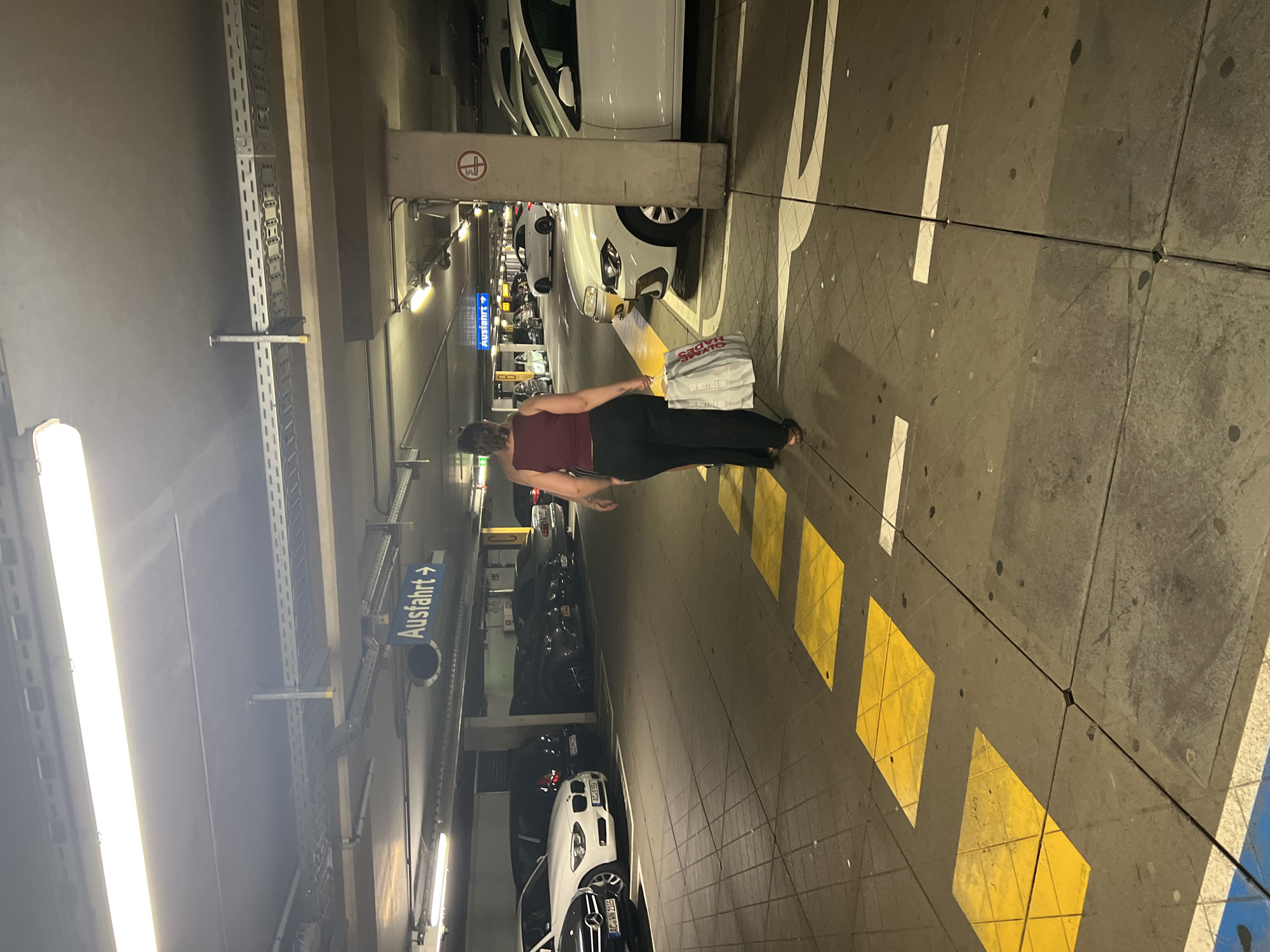 A person walks away from the camera in a dimly lit underground parking garage in Aachen, Germany. The person is wearing a dark red tank top and black pants, and is carrying a large white shopping bag. They are walking on a gray concrete floor with yellow and white painted lines and squares. Several cars are parked on either side of the aisle. Overhead, fluorescent lights illuminate the scene, and various pipes and conduits are visible along the ceiling. A blue sign with white text reading "Ausfahrt" and an arrow pointing to the right is mounted on the ceiling.