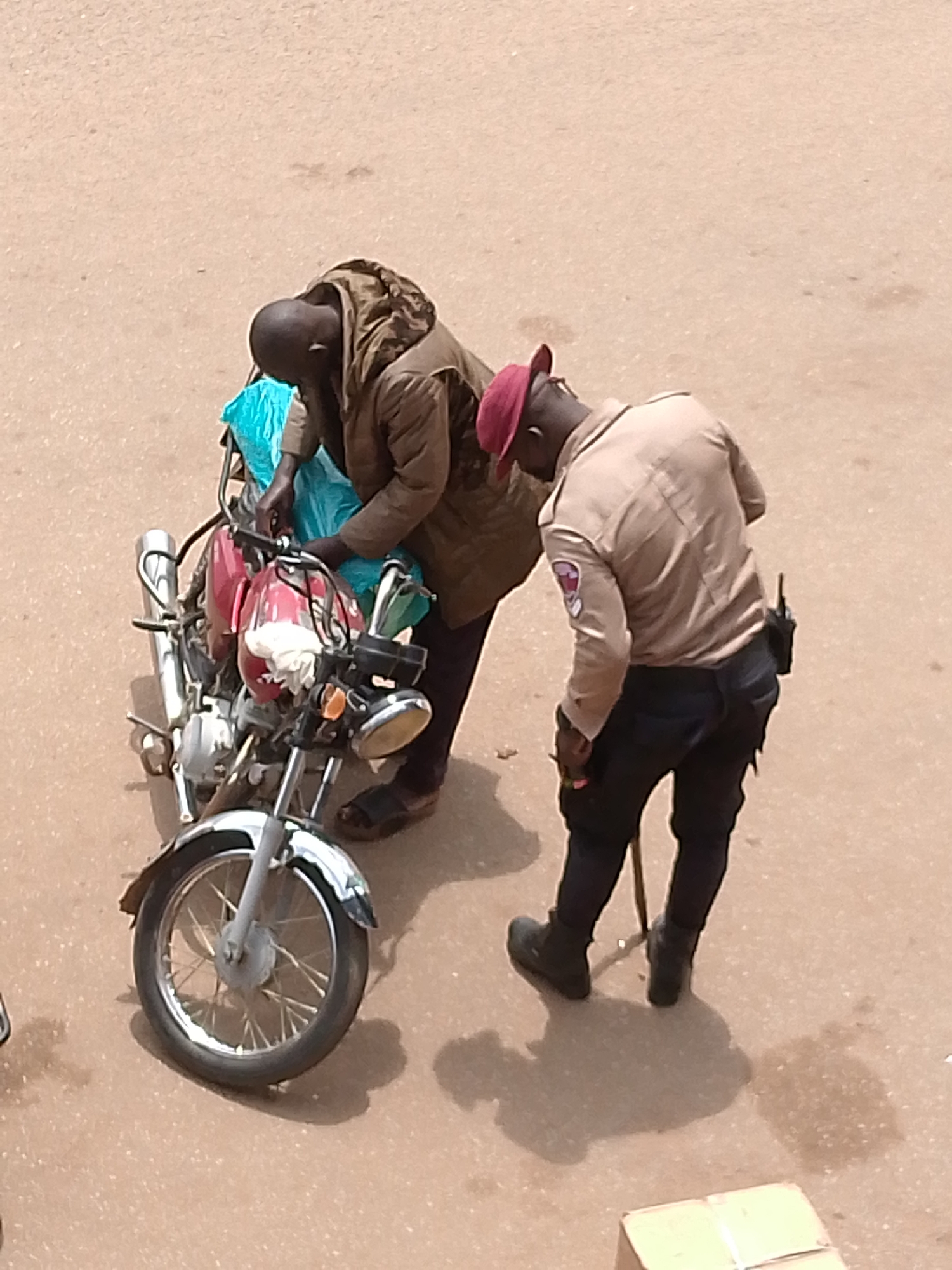 Two individuals are interacting next to a parked motorcycle on a paved surface, likely a street, in Bukuru, Nigeria. The individual on the left, wearing a brown hooded jacket and dark pants, is leaning over the motorcycle, seemingly attending to items wrapped in a blue plastic bag. Their head is down, obscuring their face. The individual on the right, dressed in a light-colored uniform shirt with an emblem on the left sleeve, dark pants, and a red beret, is bending down towards the motorcycle. This individual has a radio or communication device attached to their belt. The motorcycle is a silver and red model with a visible front wheel, handlebars, and a rear section with a silver exhaust pipe. The ground appears to be a light brown asphalt or concrete surface, with some scattered debris or discoloration. The overall lighting suggests it is daytime.