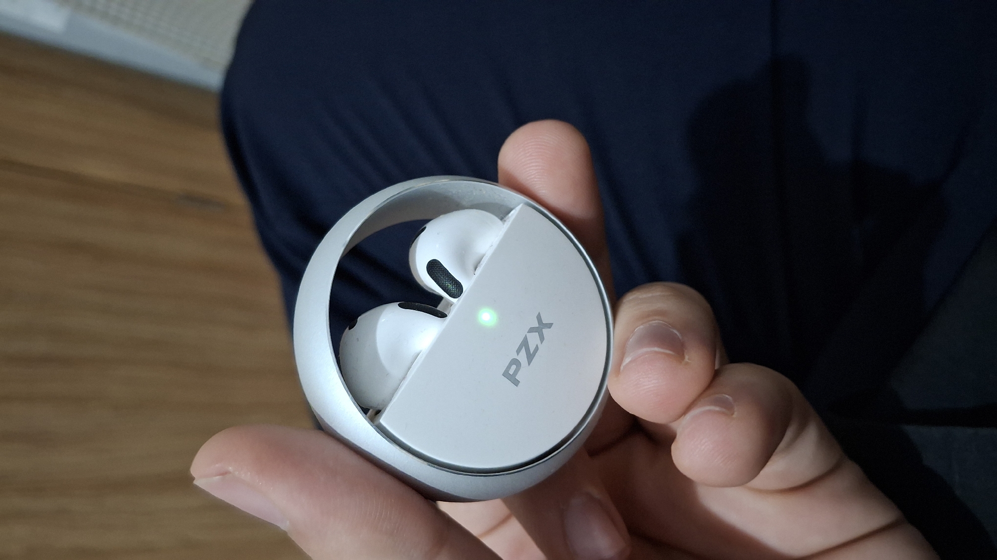 A close-up shot captures a person's hand holding a white, circular wireless earbud charging case with a silver rim. Inside the case, two white earbuds are visible. The case has a light green indicator light illuminated and the brand name "PZX" is printed on the front in gray lettering. The background is out of focus, showing a dark blue fabric surface and a wooden texture. The image does not contain any information that can be used to determine its location in the city of Nesebar, Bulgaria.