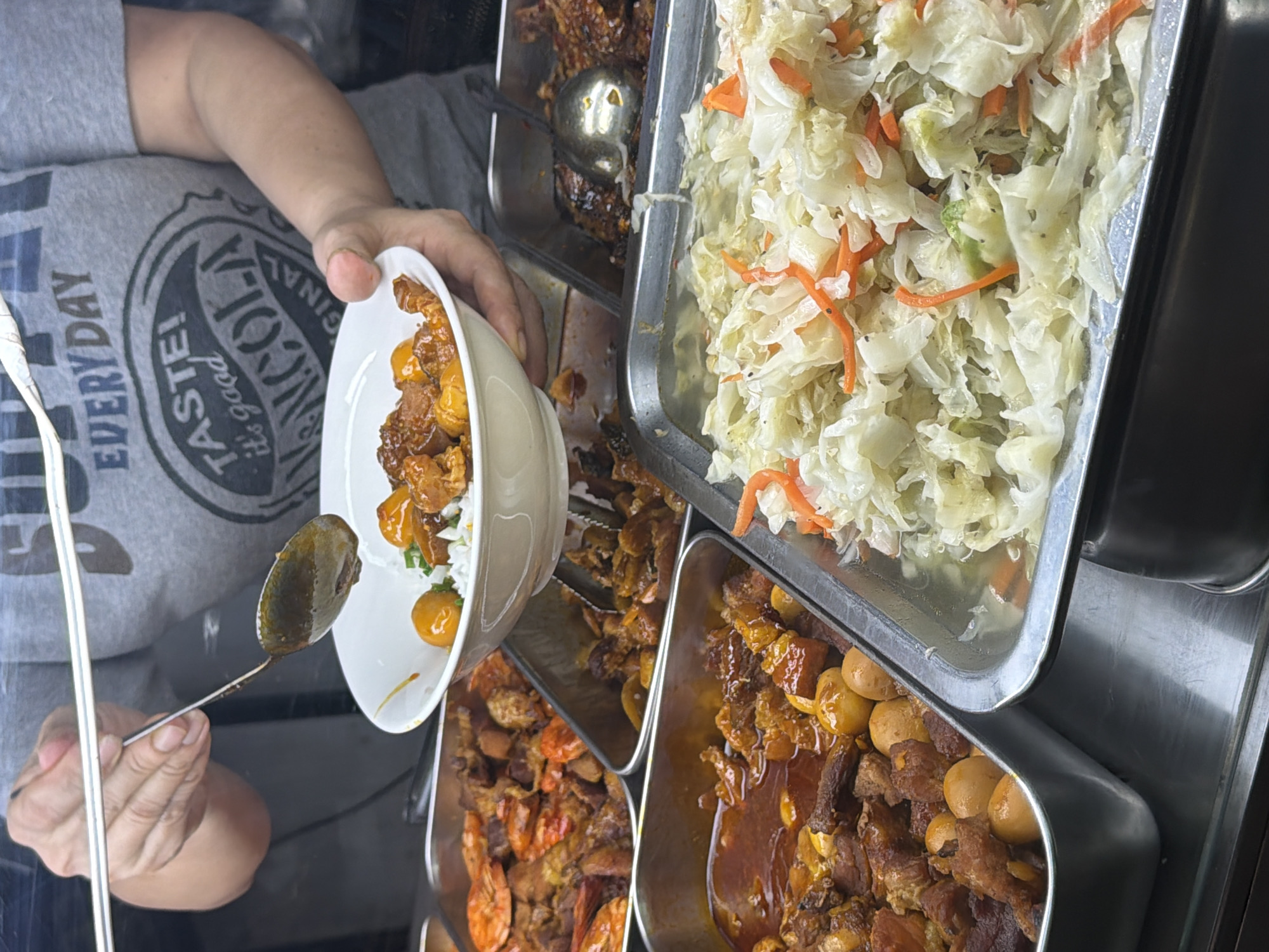 A person wearing a gray t-shirt with a graphic print is serving food from a buffet at a food stall, possibly in Nha Trang Ward, Vietnam. The t-shirt has the words "EVERY DAY" and "TASTE! it's good N.COLA GINAL" printed on it. The person is holding a white bowl with rice and what appears to be braised meat and eggs. A spoon is suspended above the bowl, with some sauce dripping from it. In the background, there are several stainless steel trays filled with various dishes, including shrimp, braised pork with eggs, and a salad of shredded cabbage and carrots. The environment suggests a casual dining or takeaway setting.