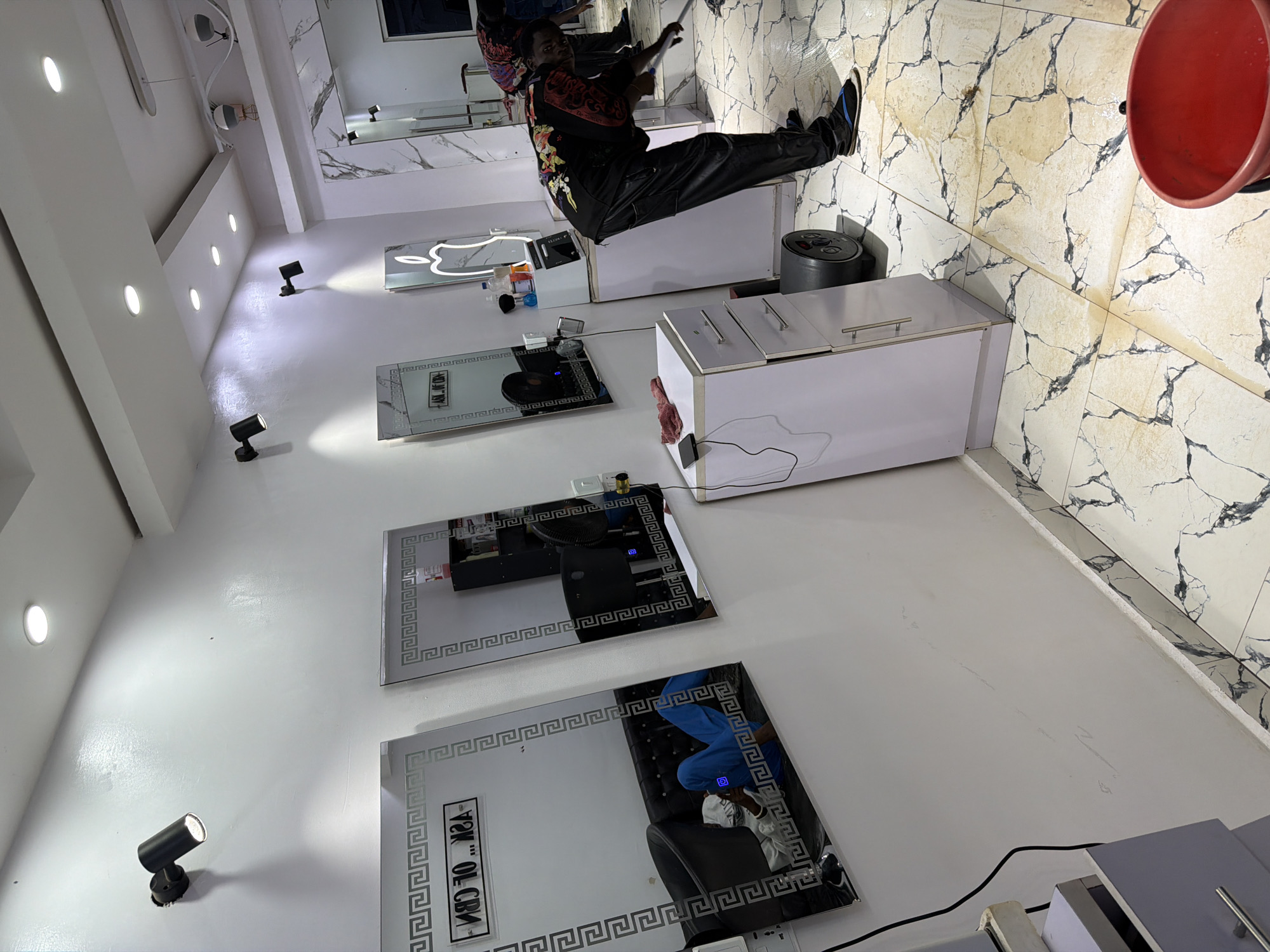 The image depicts the interior of a brightly lit salon or barbershop, likely located in Bukuru, Nigeria. The walls are painted white, and the floor is covered in white tiles with black veining. Several mirrors with decorative borders are mounted on the wall. In the foreground, a person is mopping the floor, which appears wet and has some brown staining, suggesting a recent cleaning or spillage. A red bucket is visible on the right side of the image, likely used for the mopping. There are salon chairs, hair dryers, and other salon equipment in the background. A person is seen reclining on a sofa in one of the mirrors, wearing blue pants and a white top. The overall impression is of an active, working salon environment.