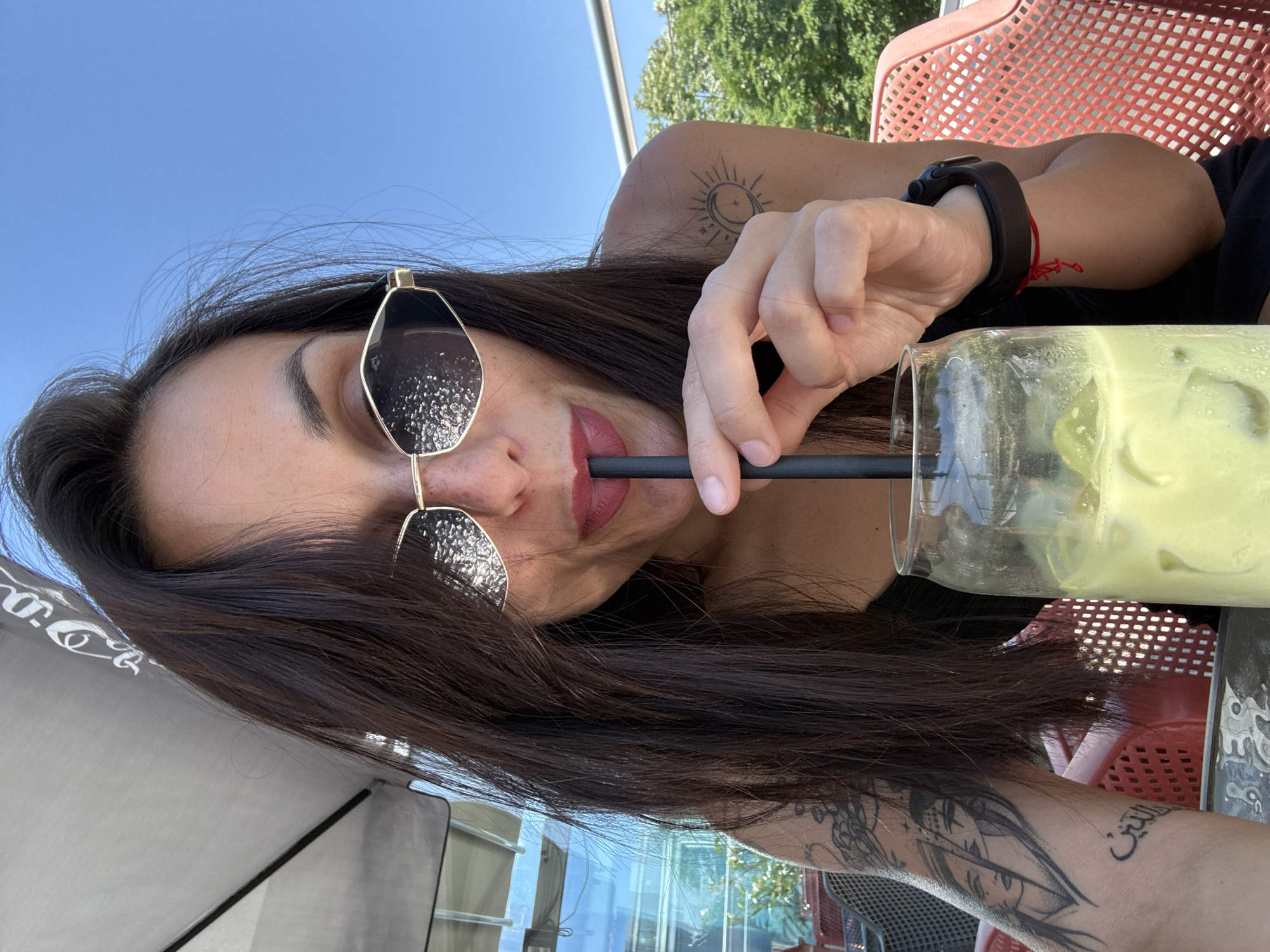 A woman with dark hair and sunglasses is drinking from a glass with a straw. She is sitting in a red plastic chair outside. She has tattoos on her arms. The sky is blue and there are trees in the background. The location is Pleven, Bulgaria.