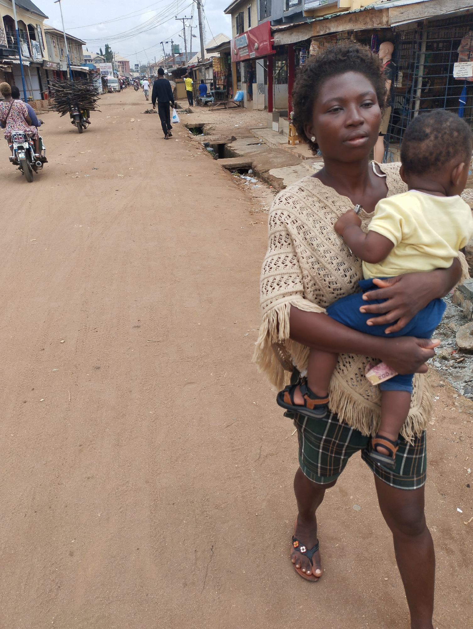 The image shows a woman holding a baby in Eziobodo, Nigeria. The woman is wearing a beige sweater with fringe, plaid shorts, and sandals. The baby is wearing a yellow shirt, blue pants, and sandals. The woman is carrying money in her hand. They are standing on a dirt road in a busy marketplace. There are other people, motorcycles, and buildings in the background. Some people on motorbikes in the background are carrying large bundles of wood.