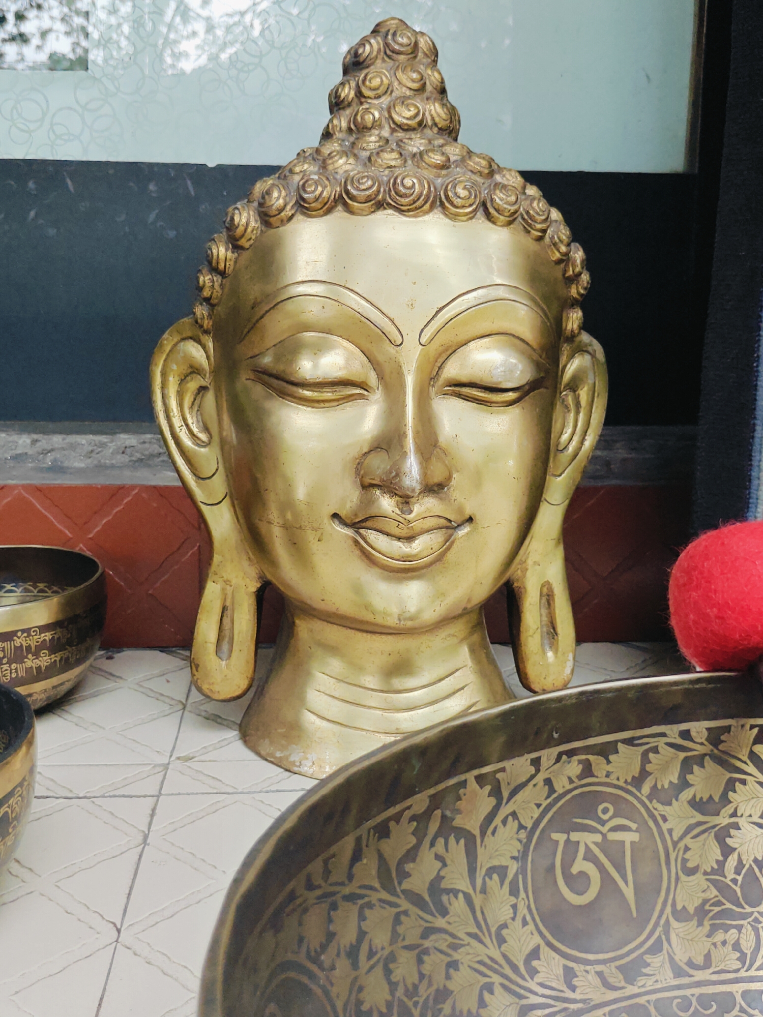 A close-up, eye-level shot captures a golden statue of a Buddha head, its eyes closed in serene meditation. The statue is positioned centrally in the frame, with its textured, coiled hair forming a prominent crown. To the left of the Buddha head, parts of two brass singing bowls are visible, one decorated with Tibetan script and floral patterns. A bright red, spherical object, possibly a pom-pom or wool ball, rests to the right of the statue. The background includes a brick-patterned wall and a darker, textured surface, hinting at an indoor setting. The lighting is soft, casting gentle highlights and shadows on the golden surfaces. The overall ambiance suggests a spiritual or artistic space. While the image prominently features cultural artifacts associated with Buddhism, it does not provide explicit information about the city of Pokhara, Nepal.