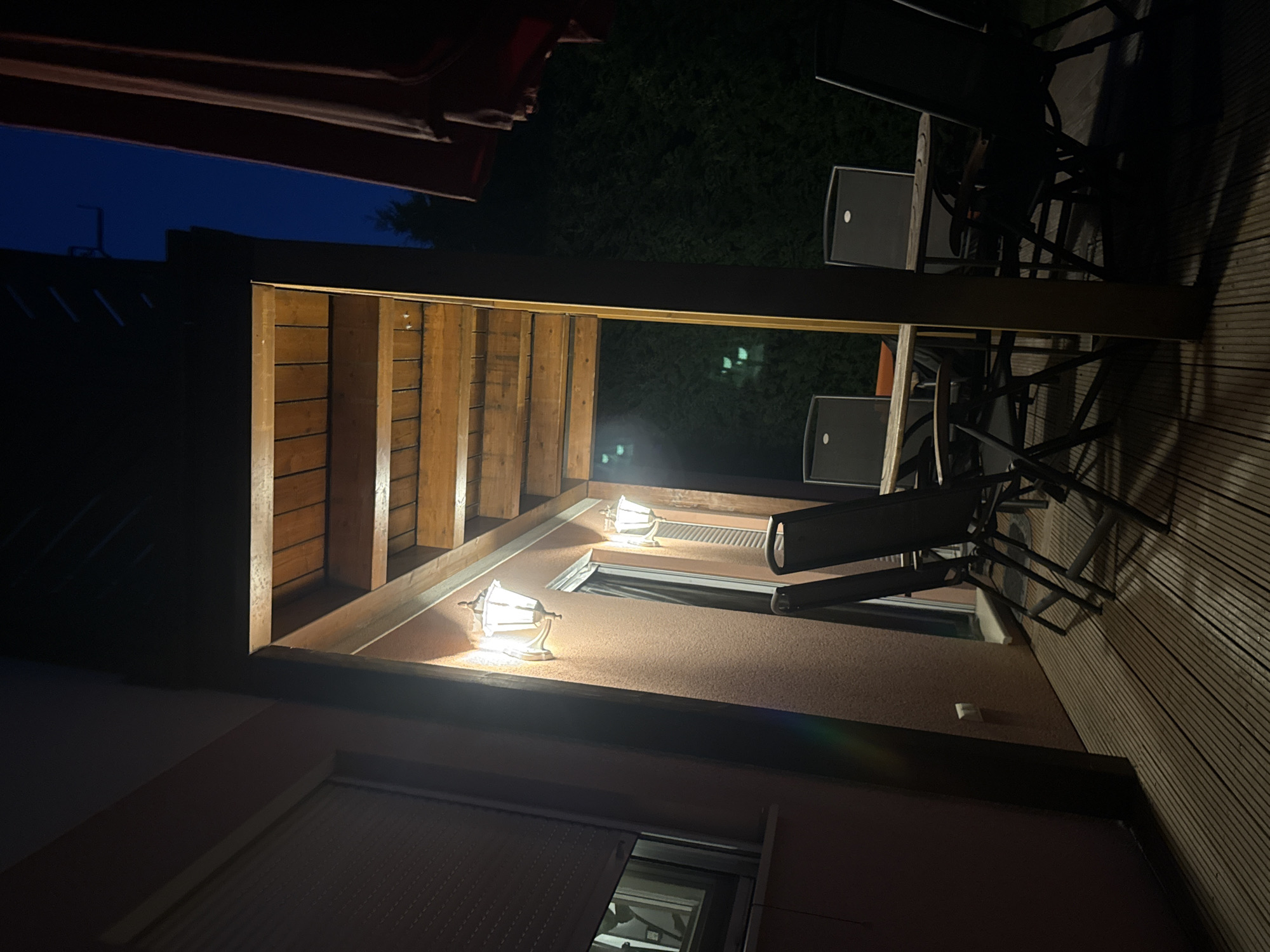 The image depicts an outdoor patio area at night, illuminated by two wall-mounted lamps casting a warm glow. A wooden pergola with exposed beams extends over the patio, which is surfaced with wooden planks. On the patio, a table with four chairs is visible, with chairs made of dark material and a wooden tabletop. To the left of the patio, a pinkish wall features a window with a rolled-down shutter and a second, smaller window to its right. The background consists of dark greenery, suggesting a yard or garden, with a deep blue twilight sky above. The scene appears to be located in Sankt Andrä-Wördern, Austria, given the context provided. No people are present in the image.