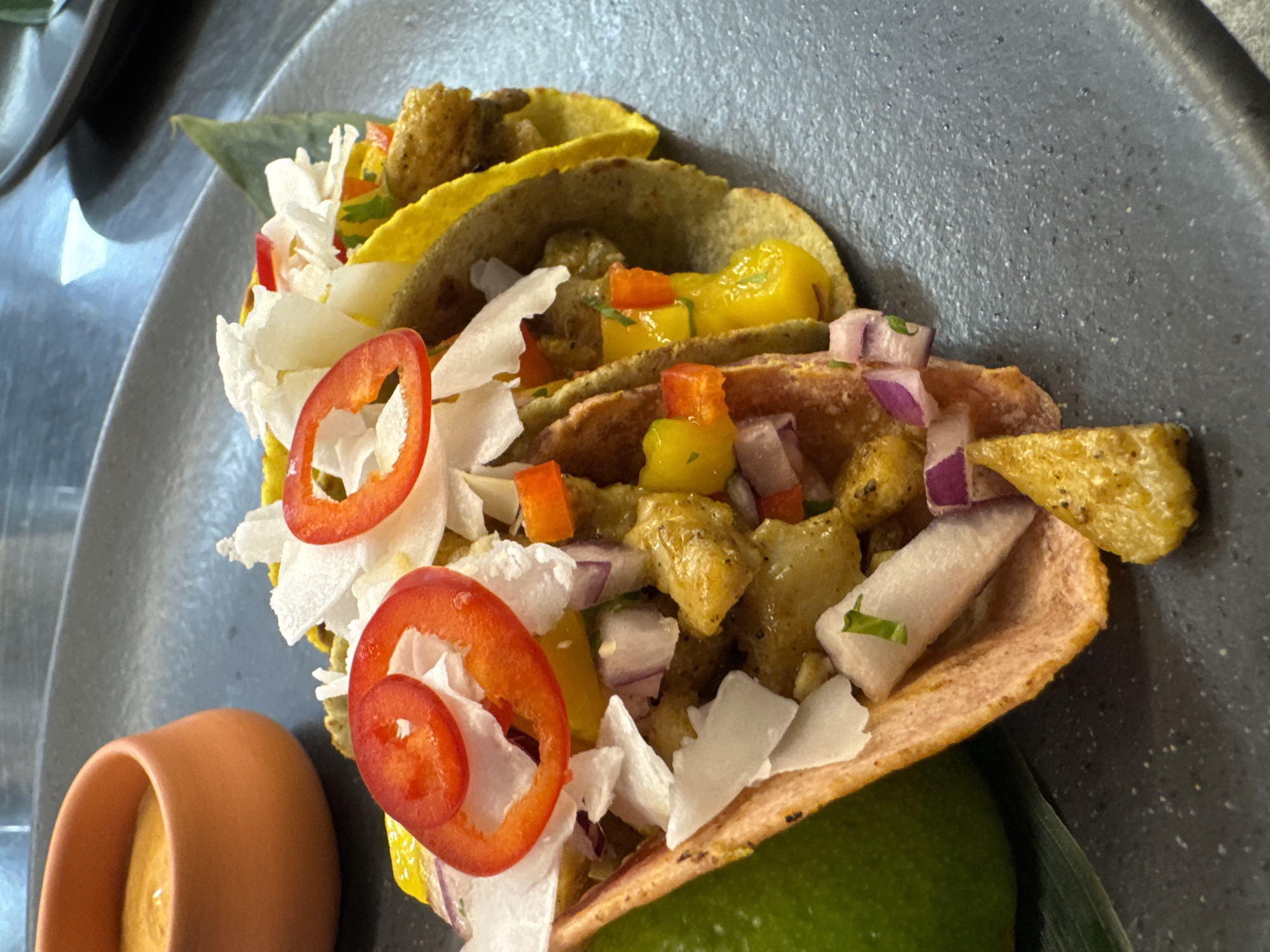 A close-up, overhead shot shows three tacos on a dark gray, textured plate. The tacos are filled with yellow and green ingredients, diced red onion, and white shreds, possibly coconut or cheese. Slices of red chili pepper are on top of the taco fillings, and a lime wedge is visible on the left side. To the left of the tacos, a small, orange, ceramic dish holds a thick, orange sauce. The background is a dark, reflective surface, possibly a table. The image appears to be taken in Sofia, Bulgaria, at a restaurant, though no specific location details are evident within the image itself.