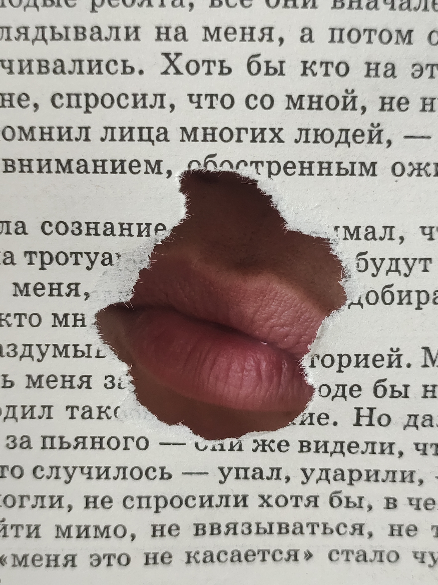 The image shows a close-up of a book page with a cutout in the shape of lips. The visible text is in Russian. The torn edges of the paper around the cutout are visible. Behind the hole, there is an image of a pair of lips. The background is out of focus, appearing to be more of the same book or another object. There is no information in the image to place it in the city of Фоки, Russia.