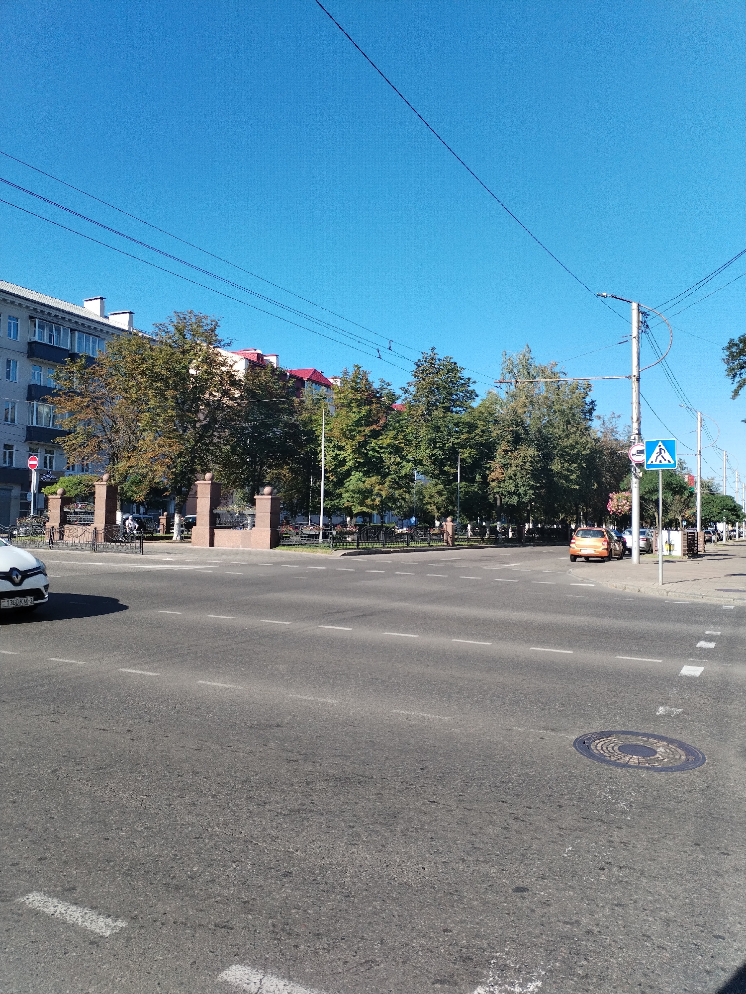 The image depicts a street scene in Homyel, Belarus, on a clear, sunny day. In the foreground, a gray asphalt road is visible with white lane markings. A white car with a license plate reading "1360 KM-3" is partially visible on the left side of the road. Further down the road, an orange car is parked on the right.
Behind the road, there is a sidewalk lined with trees and a decorative entranceway with stone pillars and metal fencing. Beyond the entrance, a building with multiple windows is visible, along with other structures with red roofs. Overhead, power lines crisscross the bright blue sky.
Several traffic signs are present, including a blue pedestrian crossing sign with a white silhouette of a person walking and a red and white circular sign with a horizontal bar. A streetlamp with visible wires is also present. In the distance, more trees and buildings can be seen, suggesting an urban environment. A manhole cover is embedded in the road on the right side of the image. A few people are present in the background, walking near the entranceway and along the sidewalk.