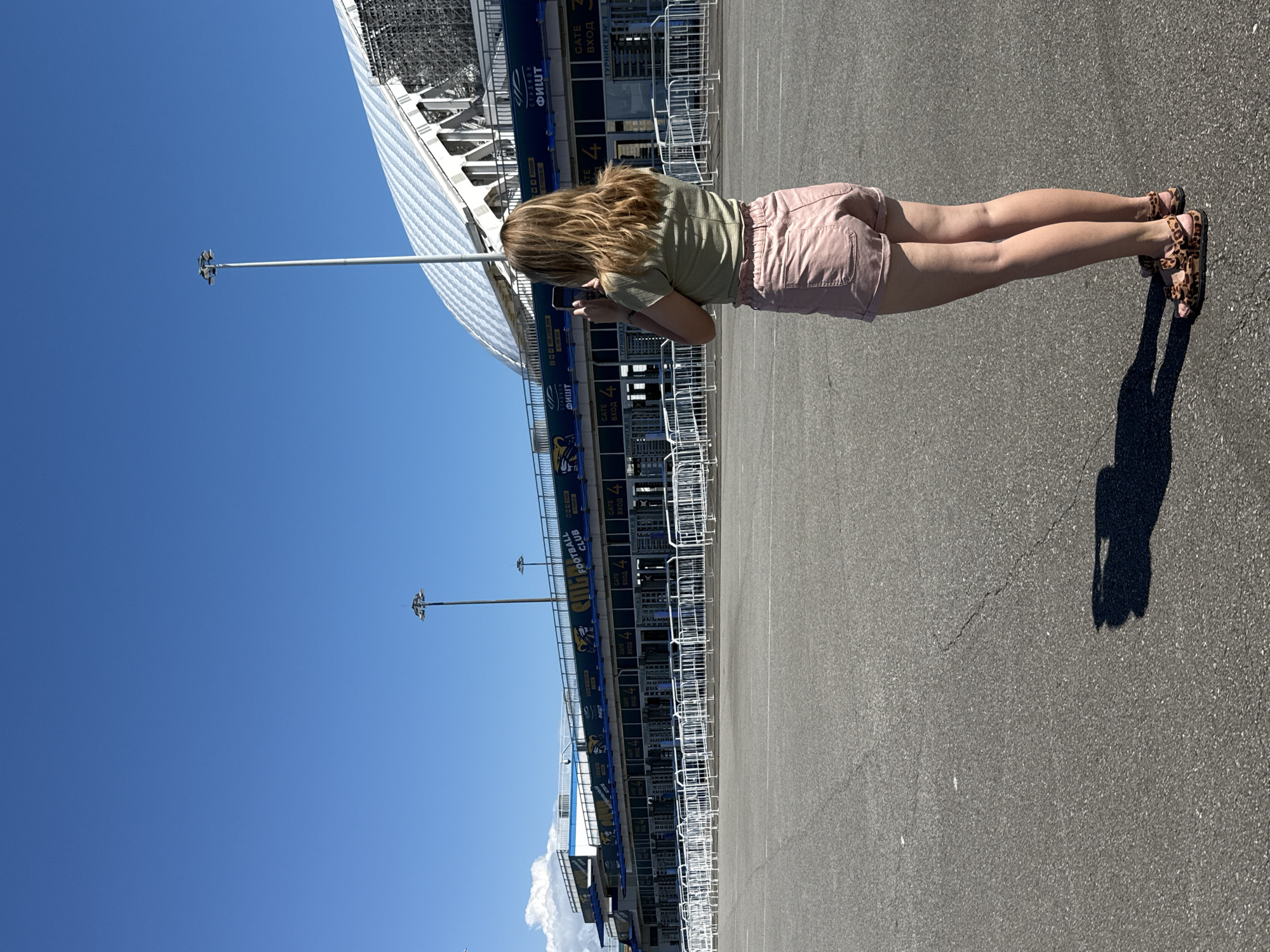 A young woman with blonde hair, wearing a pale green t-shirt and pink shorts, stands facing away from the camera, holding a smartphone up to take a picture. She is on an asphalt surface in front of a stadium entrance. The stadium has a blue and yellow facade with the words "STADIUM FISHT" and "FOOTBALL CLUB" visible, along with multiple entry gates and turnstiles marked with "GATE" and "ВХОД" (entrance in Russian). White metal barriers line the area in front of the gates. In the background, the distinctive curved white roof of the Fisht Olympic Stadium is visible against a clear blue sky. The overall setting appears to be the city of Sirius, Russia, given the prominence of the Fisht Stadium, a well-known landmark in the region. The lighting suggests a bright, sunny day, casting a shadow of the woman and her phone onto the ground in front of her.