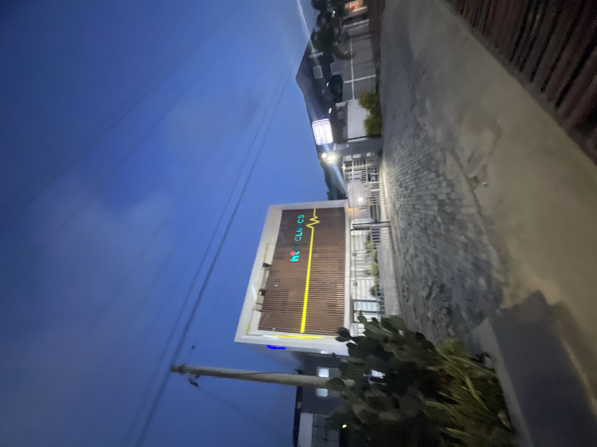 The image shows a building identified as "KC Clinics" in Ilado, Nigeria, taken at dusk or night. The building has a modern design with a primarily brown facade and a prominent yellow and green medical symbol. A tall pole is located to the left of the building, running through the bottom of the frame. The sky is a deep blue. A street or walkway runs to the right of the building, and there is some vegetation in the foreground. Other buildings are visible in the background.