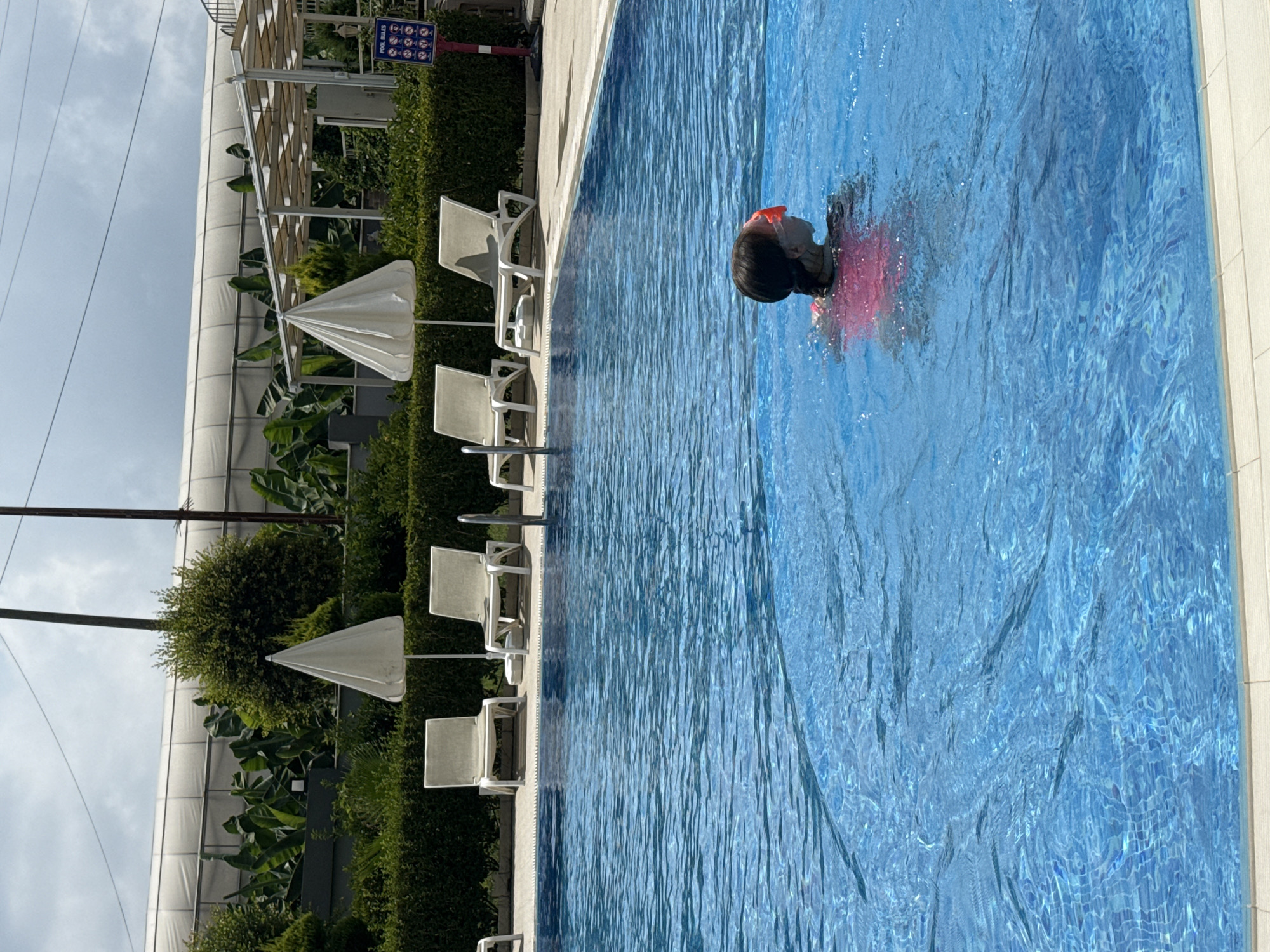 The image shows a young child wearing goggles and a pink swimsuit swimming in a bright blue swimming pool. The pool has tiled flooring visible beneath the water. In the background, there are several lounge chairs and white umbrellas lined up on the pool deck. Beyond the pool area, lush green hedges and trees are visible. In the far background, a large white structure, possibly a greenhouse or covered facility, is present, along with some power lines overhead. A red sign with white text and icons, likely displaying pool rules, is mounted on a post to the right of the image. The overall scene suggests a resort or hotel with a swimming pool in Alanya, Turkey.