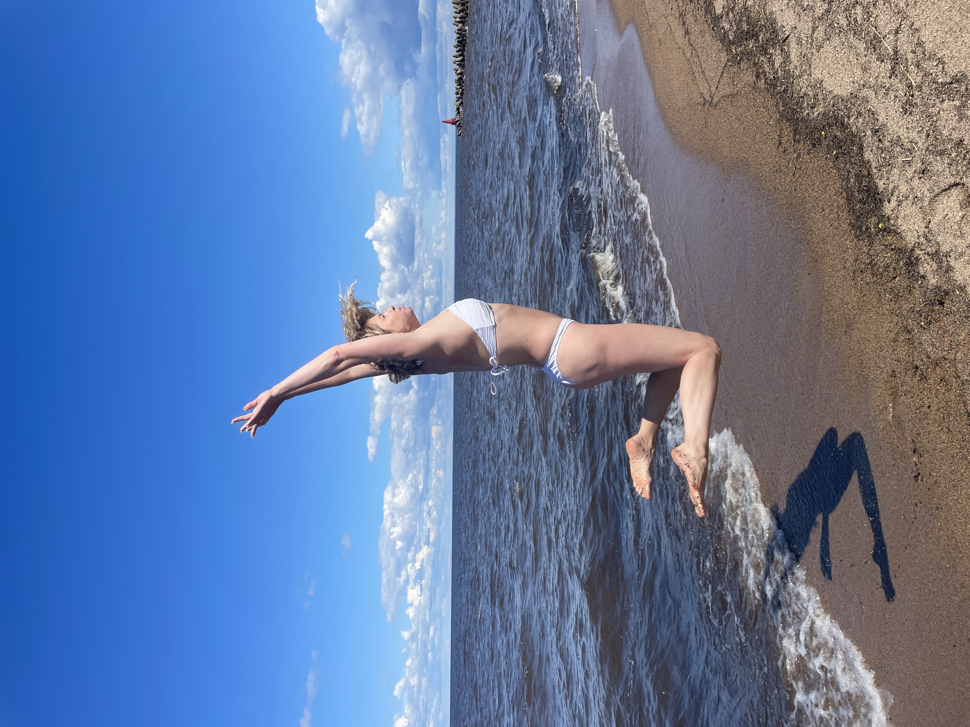 A full-body shot shows a woman in a white bikini jumping on a sandy beach next to the dark blue water of a large lake or sea. The sky is bright blue with scattered white clouds. The woman is in mid-air, with her arms raised above her head and her legs bent. Her body is in profile, facing to the right. The water has small waves crashing on the shore. In the distance, there is a breakwater made of large rocks and a red navigational marker. The sand is a light brown color and appears to be damp near the water's edge. The sun is casting a shadow of the woman on the sand. The scene is likely located in Belozersk, Russia, given the context provided by the prompt, which suggests it's a sunny day on the shore of a body of water.