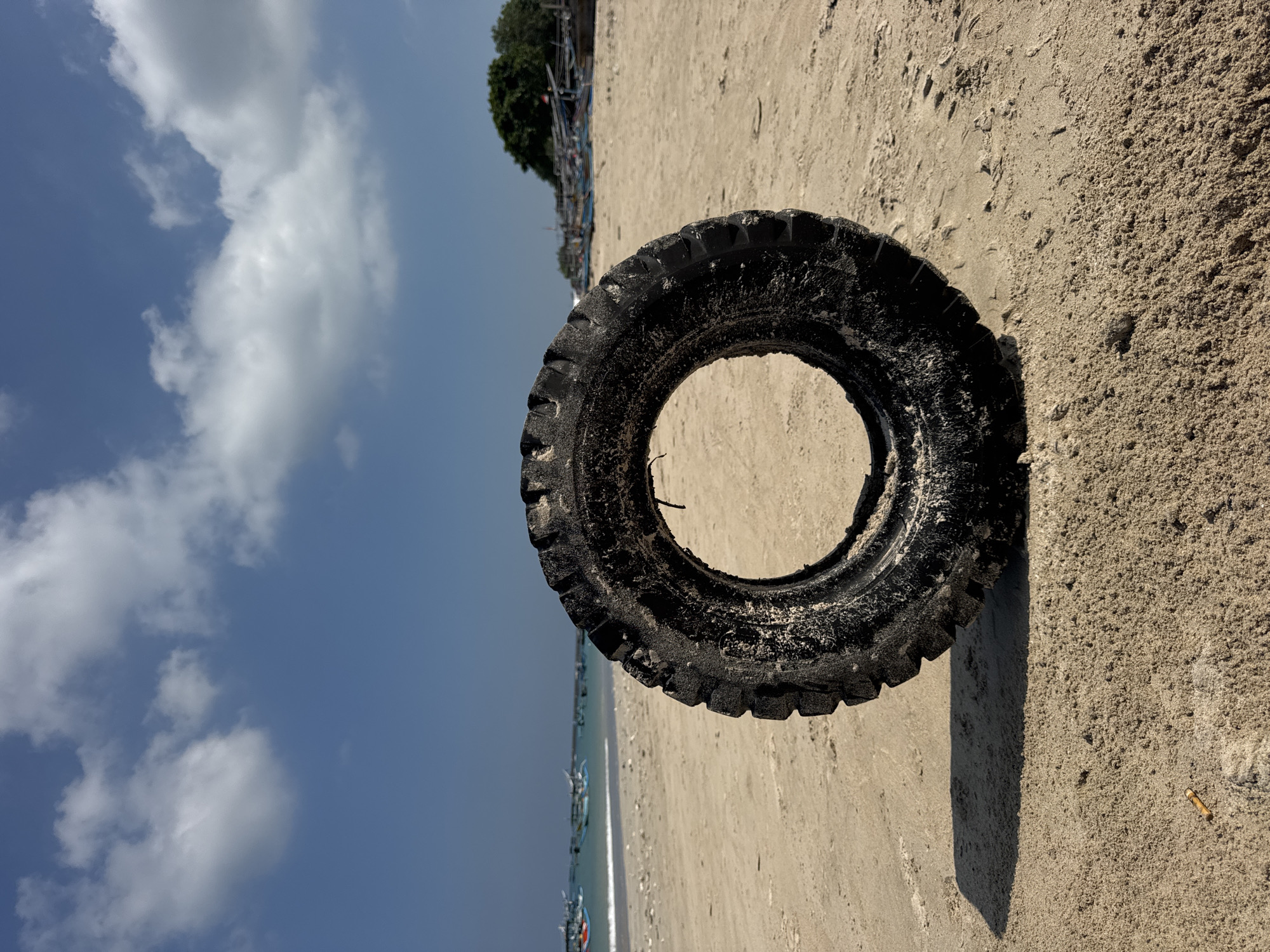 This vertically oriented image captures a coastal scene, likely in Kuta, Indonesia, dominated by a large, well-worn, black tire resting on a sandy beach. The tire, appearing to be from a heavy vehicle or farm equipment due to its deep treads, is encrusted with sand, highlighting its grimy texture. Its dark form casts a distinct shadow on the light-brown sand to its right. A cigarette butt is visible on the sand near the shadow.
Beyond the tire, the scene extends to a tranquil body of water, possibly the ocean, with a gentle wave line visible along the shore. Numerous small, traditional fishing boats with colorful hulls are either anchored or docked in the shallow waters, stretching into the distance. Further in the background, a line of dense green trees or foliage marks the edge of the land.
The upper portion of the frame features a bright blue sky, adorned with scattered, fluffy white cumulus clouds, suggesting a clear and sunny day. The sunlight is strong, creating clear shadows. No specific human activity is depicted, but the boats and the tire suggest a working beach environment. The image is devoid of any visible text. The composition emphasizes the contrast between the natural elements of sand, sea, and sky, and the man-made object of the tire.