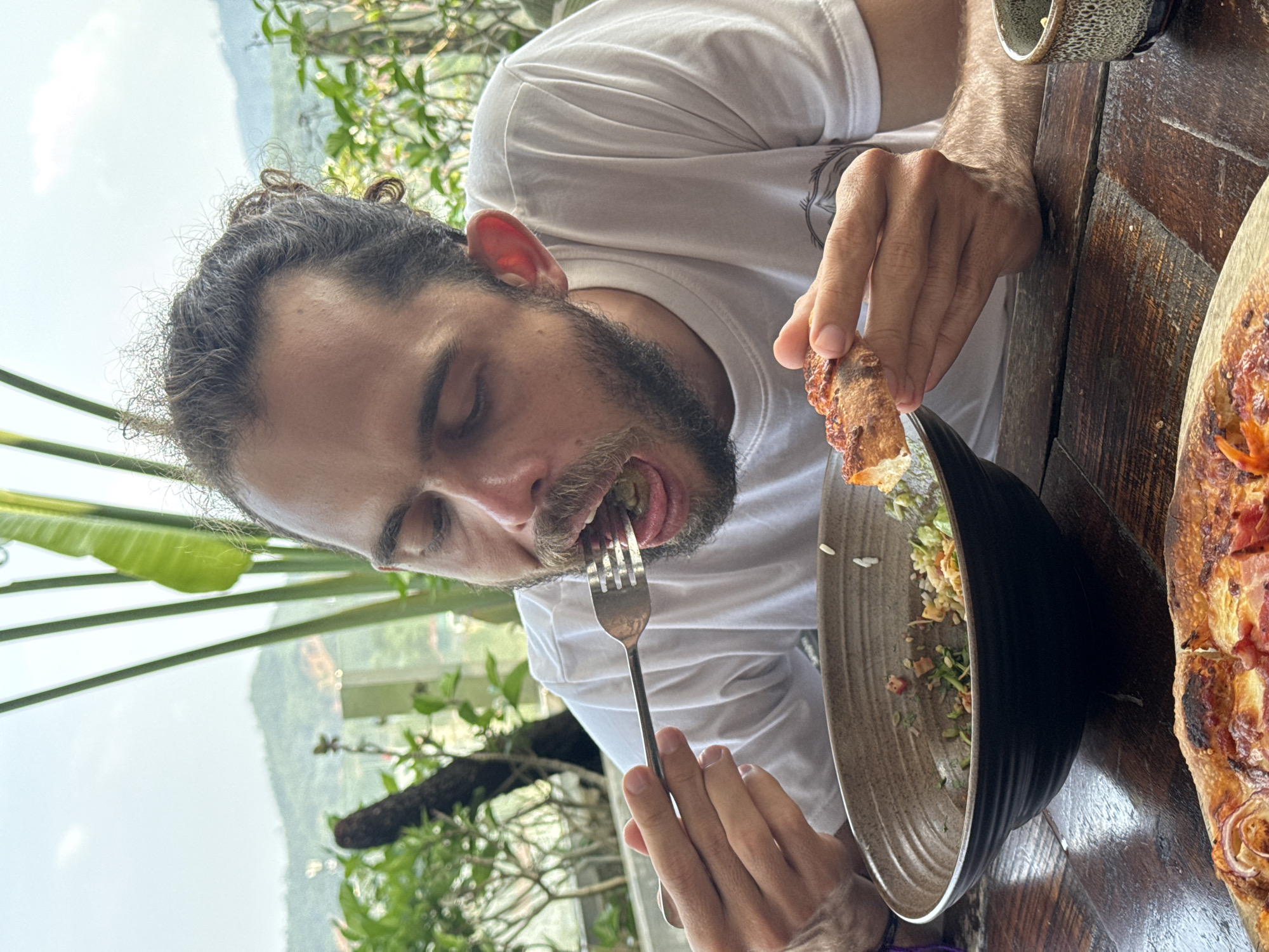 A man with dark, curly hair and a beard, wearing a white t-shirt, is depicted in the act of eating at a rustic wooden table. His head is tilted downwards, and his eyes are closed or deeply focused on his meal. A fork is visible entering his open mouth, carrying food from a dark, textured bowl that contains a partially eaten salad or rice dish. His left hand holds a small piece of crusty food, possibly a piece of pizza or bread, near his face.
The table features a long, rectangular pizza with toppings that appear to include ham or bacon and red onions. A small, patterned ceramic cup or bowl is also visible on the wooden surface.
The setting is an outdoor, open-air environment with abundant green foliage and trees serving as the immediate backdrop. Beyond the close-up greenery, a bright, expansive sky stretches over distant mountains or hills, suggesting a scenic vista. The soft, natural lighting indicates it is daytime. The scene conveys a moment of focused enjoyment of a meal in a natural and picturesque location, consistent with the context of Hà Giang 1 Ward, Vietnam. There is no discernible text in the image.