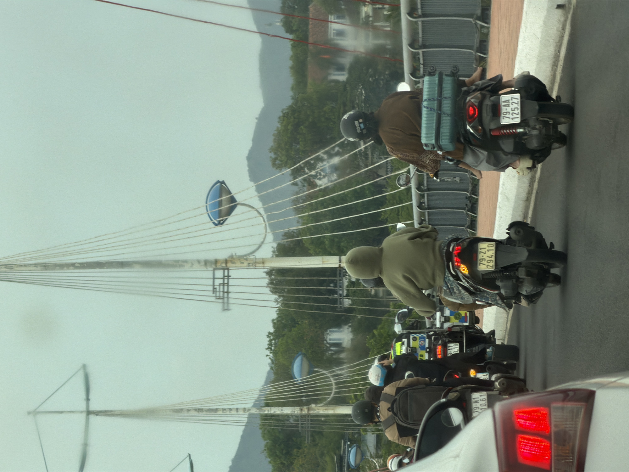 The image shows a bridge in Nha Trang Ward, Vietnam, with a convoy of motorcycles traveling on it. The bridge is supported by cables attached to tall, blue-topped towers. A white car is partially visible in the foreground, with its red taillights illuminated. The motorcycles are carrying passengers and some luggage, as evidenced by the blue suitcase strapped to the back of one. The sky is overcast. License plates are visible on the motorcycles, reading "79-AA 125.27", "79-Z1 294.10", "9-A1 3.18 004018", and "79-N1 10.61". The motorcycles appear to be in motion. Hills are visible in the background.