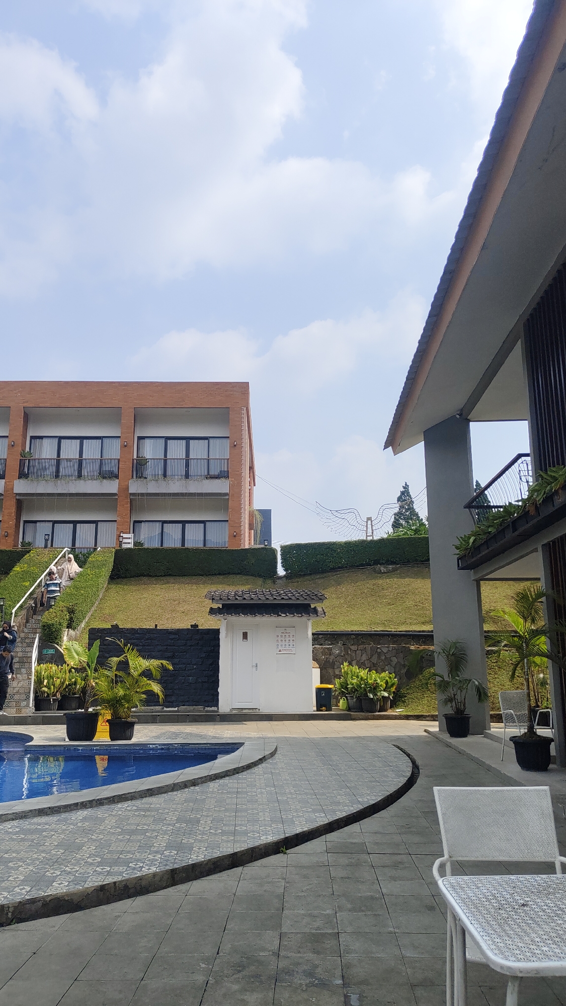 This is a vertical, eye-level, full outdoor shot of a resort area in Cisarua, Indonesia, on a bright, partly cloudy day. The foreground features a paved area with a patterned gray tile surface and a white metal table with chairs. To the left, a section of a blue swimming pool is visible. In the midground, a white building with a dark tiled roof stands next to a black brick wall. There are several potted plants, including palm trees, around the pool and near the white building. Behind the building, a grassy slope is covered with manicured hedges.
On the left side of the midground, a multi-story brick building with dark-framed windows and balconies is situated. People are seen on a staircase leading up to this building, and a few more are visible on the upper balconies. Further in the background, a large, stylized wire sculpture resembling wings is perched on top of the slope. To the right of the midground, a modern building with a gray concrete pillar and a dark-slatted facade is partially visible, with an open walkway and stairs. Potted plants and a white metal chair are also present in this area. The overall environment suggests a well-maintained resort with amenities like a pool and various architectural structures.