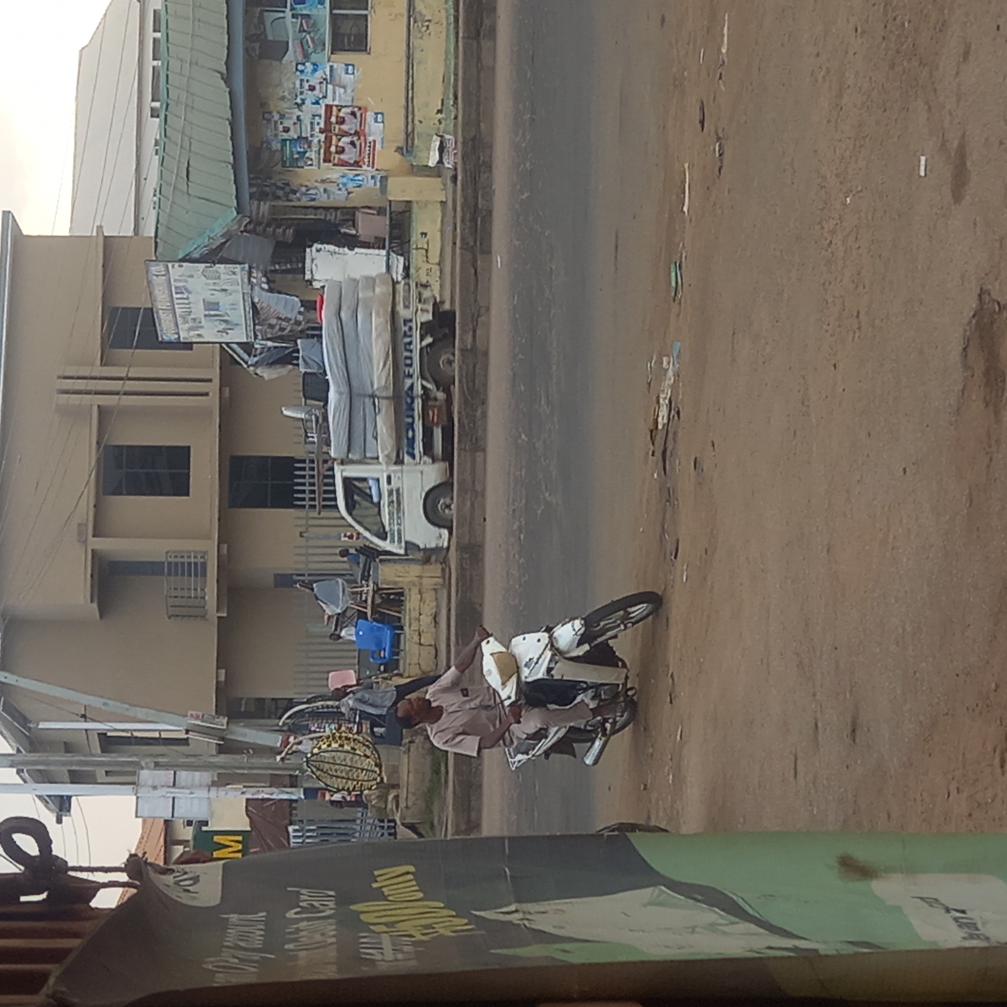 The image shows a street scene in what appears to be Dadi Riba, Nigeria. A man on a white motorcycle is in the foreground. A white truck loaded with mattresses, labeled "OSUKA FOAM," is parked along the side of the road near a two-story building. The building has multiple windows and what appears to be an advertising sign or poster on its side. Other objects, including chairs and miscellaneous items, are visible near the building. The road is unpaved. A blurred advertisement banner is partially in the foreground.