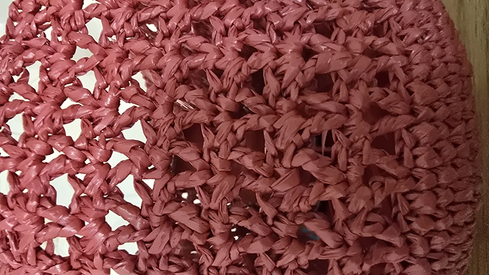 A close-up, slightly angled shot shows a dense, crocheted or woven material in a reddish-pink color. The material appears to be made from thin, twisted strands, creating an open-knit, mesh-like pattern. There are visible gaps in the weave, allowing light to pass through. The texture is rough and somewhat irregular, with individual strands clearly discernible. The lighting casts subtle shadows, highlighting the three-dimensional nature of the weave. The bottom of the image shows a hint of a surface beneath the material, possibly wood or a similar textured material. There are no people, animals, or recognizable landmarks present. The scene is entirely focused on the texture and pattern of the woven material.