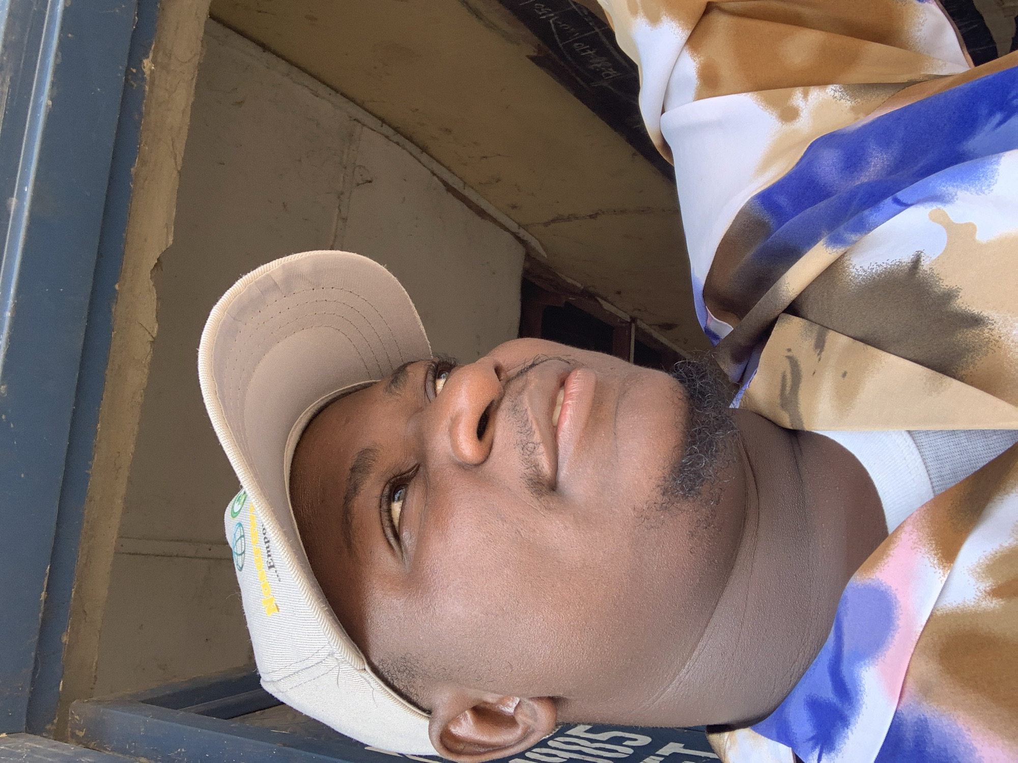 The image is a medium-close-up selfie of a dark-skinned man wearing a light-colored baseball cap and a white t-shirt under a colorful shirt with a tie-dye pattern of brown, blue, and pink. He has a goatee and is looking to his right, with his head tilted slightly upwards. The background appears to be outdoors, possibly in a semi-urban or rural setting. There is a dark blue metal structure above him, and a wall with some writing or markings on it to his right. The lighting suggests it is daytime, with sunlight casting shadows. The image does not provide specific details to pinpoint the location within Akwanga, Nigeria, but it suggests a scene that could be found in various parts of the town, such as near a market, a construction site, or an informal gathering place.