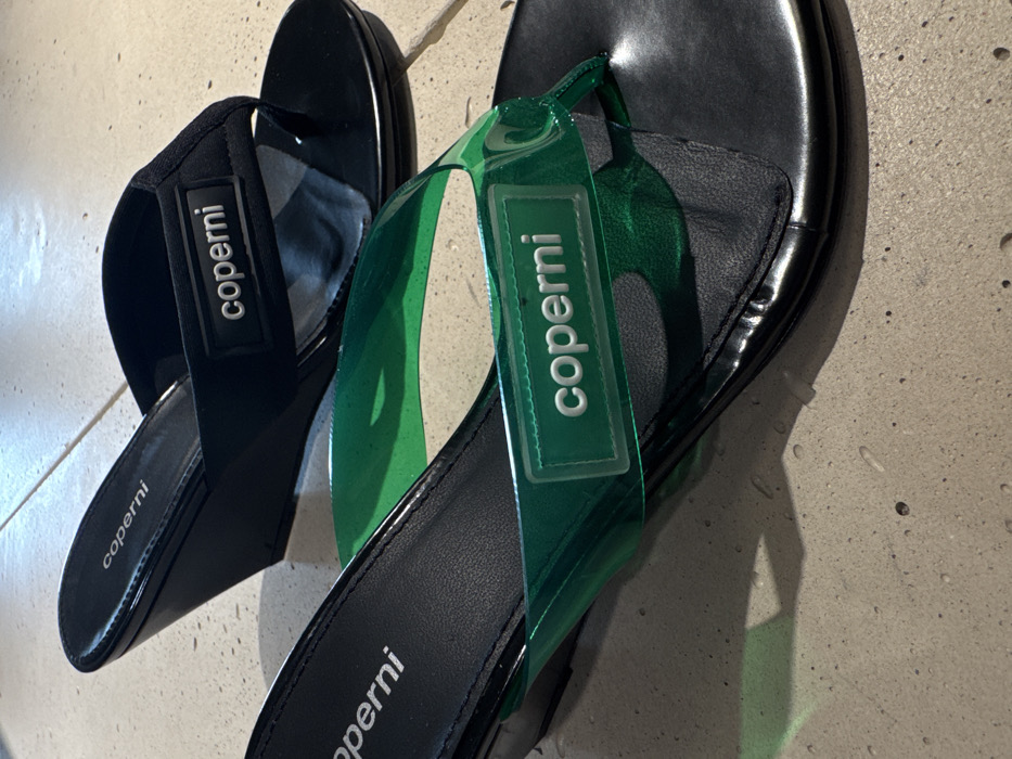 The image displays two pairs of Coperni branded wedge sandals. In the foreground, one black sandal features a translucent emerald green plastic upper strap with "coperni" in white lettering on a clear rectangular label, accompanied by a black wedge heel. Positioned behind it, a second black sandal, also with a black wedge heel, has a black fabric upper strap with "coperni" in white lettering on a black rectangular label. Both sandals are thong-style. They are situated on a light gray, textured surface, consistent with polished concrete or large-format tile, under bright illumination. No individuals are present, and there are no discernible landmarks or specific architectural features within the image to identify a precise location within Paris, France.