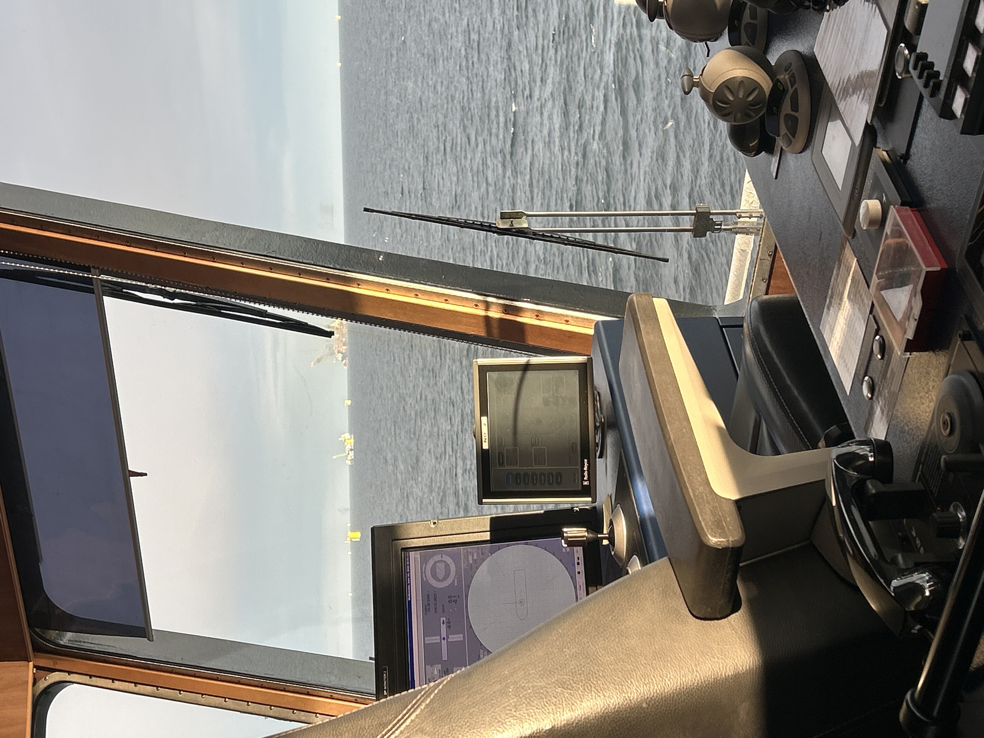 The image depicts the interior of a ship's bridge, offering a view of the sea and distant structures. In the foreground, a dark-colored captain's chair with a textured surface is prominently featured on the left. Adjacent to the chair are two computer monitors displaying various navigational data. The monitor on the left shows a circular display with lines and numbers, possibly indicating a radar or sonar system. The monitor on the right, labeled "NO. 2," presents a grid of numbers and text, with "Rolls Royce" visible at the bottom.
The bridge console features several controls and instruments. A dark-colored lever with a chrome accent is located on the left side of the console. A small red-lidded plastic box sits on a black surface. Various buttons and knobs are visible on the console. A windshield wiper mechanism is mounted on the exterior of the bridge window, angled to the right.
The expansive window offers a view of a calm, grey sea under an overcast sky. In the distance, several offshore structures are visible, including what appear to be oil rigs and buoys, some of which are yellow. The horizon is clearly defined between the sea and the sky. The overall lighting suggests daylight, with some sunlight catching the edge of the chair.