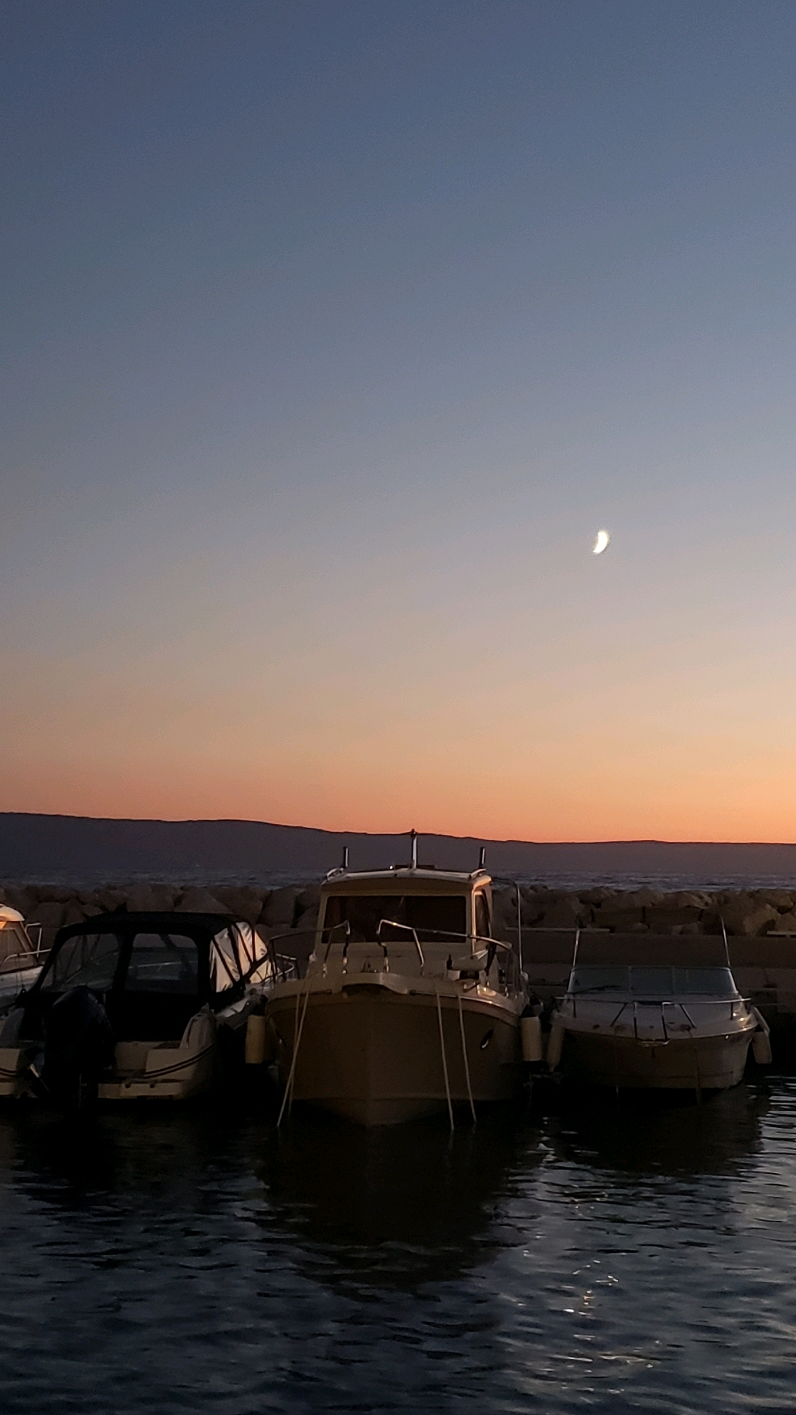 In Općina Tučepi, Croatia, a twilight scene unfolds at a harbor. The sky transitions from a deep blue overhead to a soft orange and pink hue near the horizon, where a distant mountain range is silhouetted. A crescent moon is visible in the upper right quadrant of the sky. Three boats are moored in the dark, rippling water, their reflections creating elongated shapes. The boats are docked alongside a stone breakwater. The boat on the far left is partially obscured, but its dark canopy and outboard motor are visible. The central boat is light-colored with a cabin and appears to be the largest. The boat on the far right is also light-colored and appears to be a smaller vessel. The overall atmosphere is tranquil and serene, characteristic of the end of the day in a coastal town.