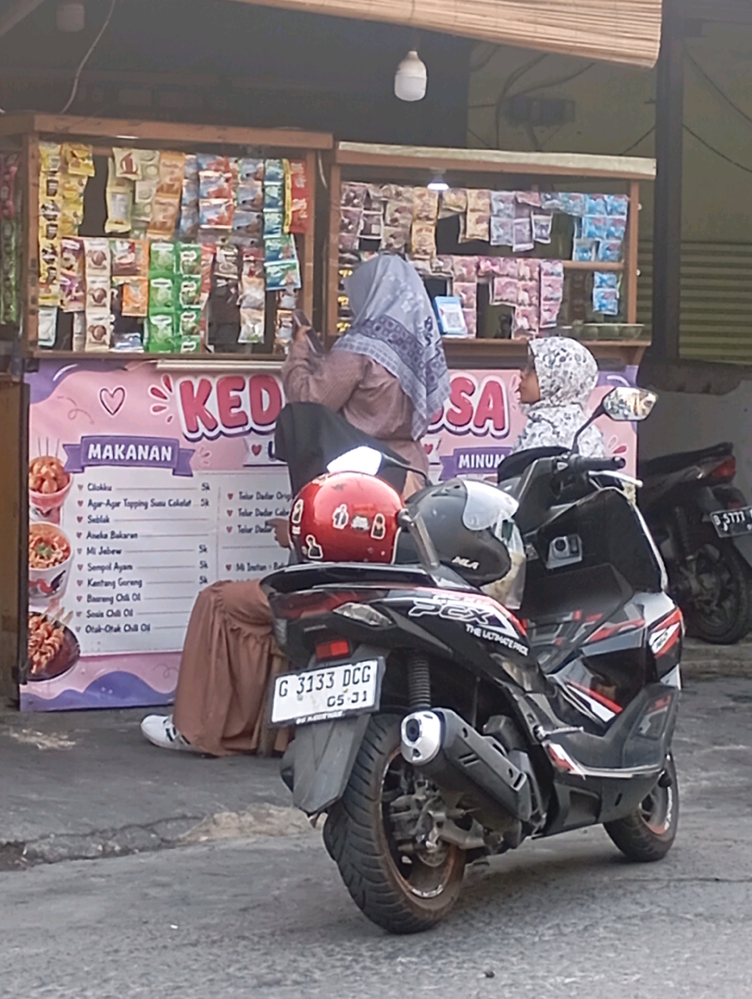 In Babelan, Indonesia, a street food stall is depicted. Two women, one in a light purple hijab and the other in a patterned white hijab, are present at the stall. The woman in the purple hijab is seated, looking at her phone, while the woman in the white hijab is standing and appears to be interacting with the stall. In the foreground, a black motorcycle with the license plate "G 3133 DCG 05-31" is parked. The stall itself displays various packaged snacks on shelves and a menu board listing food items like "Cireng," "Aneka Bakaran," and "Otak-Otak Chill Oil." The overall environment suggests a casual outdoor setting in a commercial area.