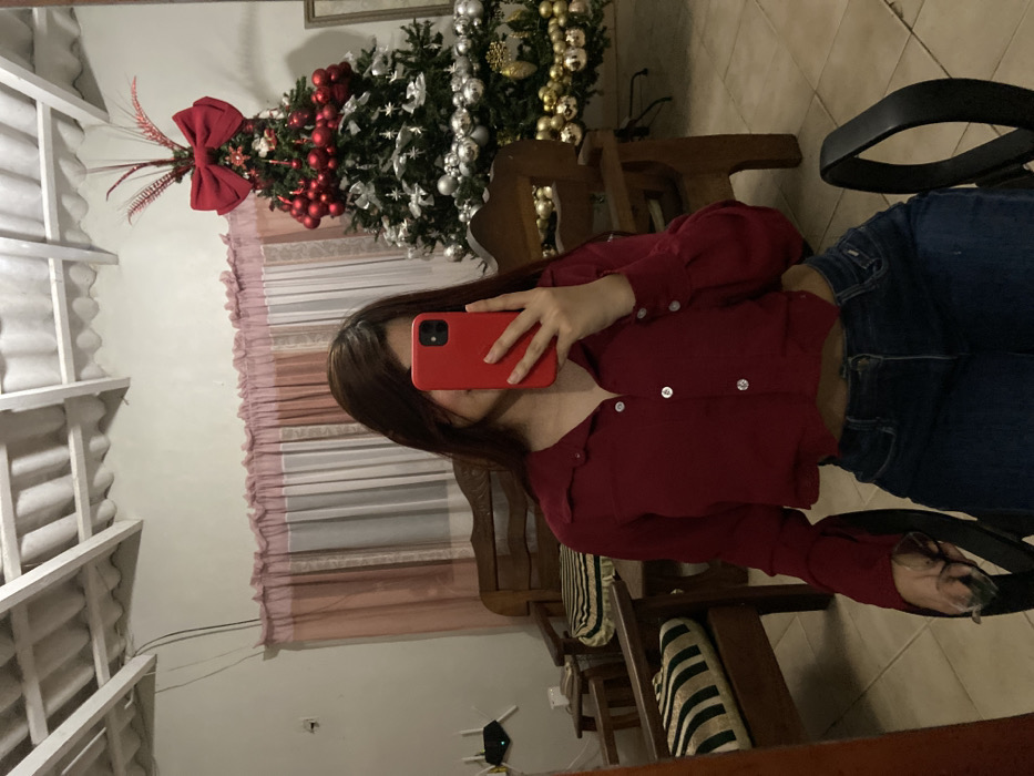 A person stands in front of a mirror, using a red phone to take a picture. She wears a red, button-down shirt and blue jeans. In the background, there is a Christmas tree decorated with silver and gold ornaments and a large red bow, visible alongside a window with white frames. Wooden chairs are also visible. The image gives the impression of a casual, festive setting within a home.