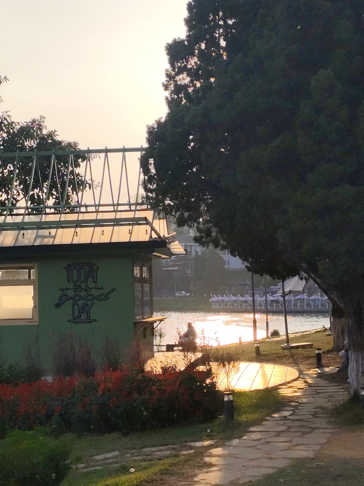 The photograph captures a tranquil scene at Xuan Huong Lake in Da Lat, Vietnam. In the foreground is a small, green building with a unique, open-air roof structure, adorned with the words "TODAY IS A good DAY". A flowerbed with red blossoms adds a touch of color at its base. To the right of the building, a stone path leads towards the lake, illuminated by the setting sun, which casts a warm glow on the path and the water. A large tree dominates the right side of the frame. In the middle distance, figures can be seen sitting near the water's edge, enjoying the peaceful atmosphere. Across the lake, there's a glimpse of buildings and a restaurant with white umbrellas, providing a backdrop to the scene. The overall impression is one of serenity and natural beauty.