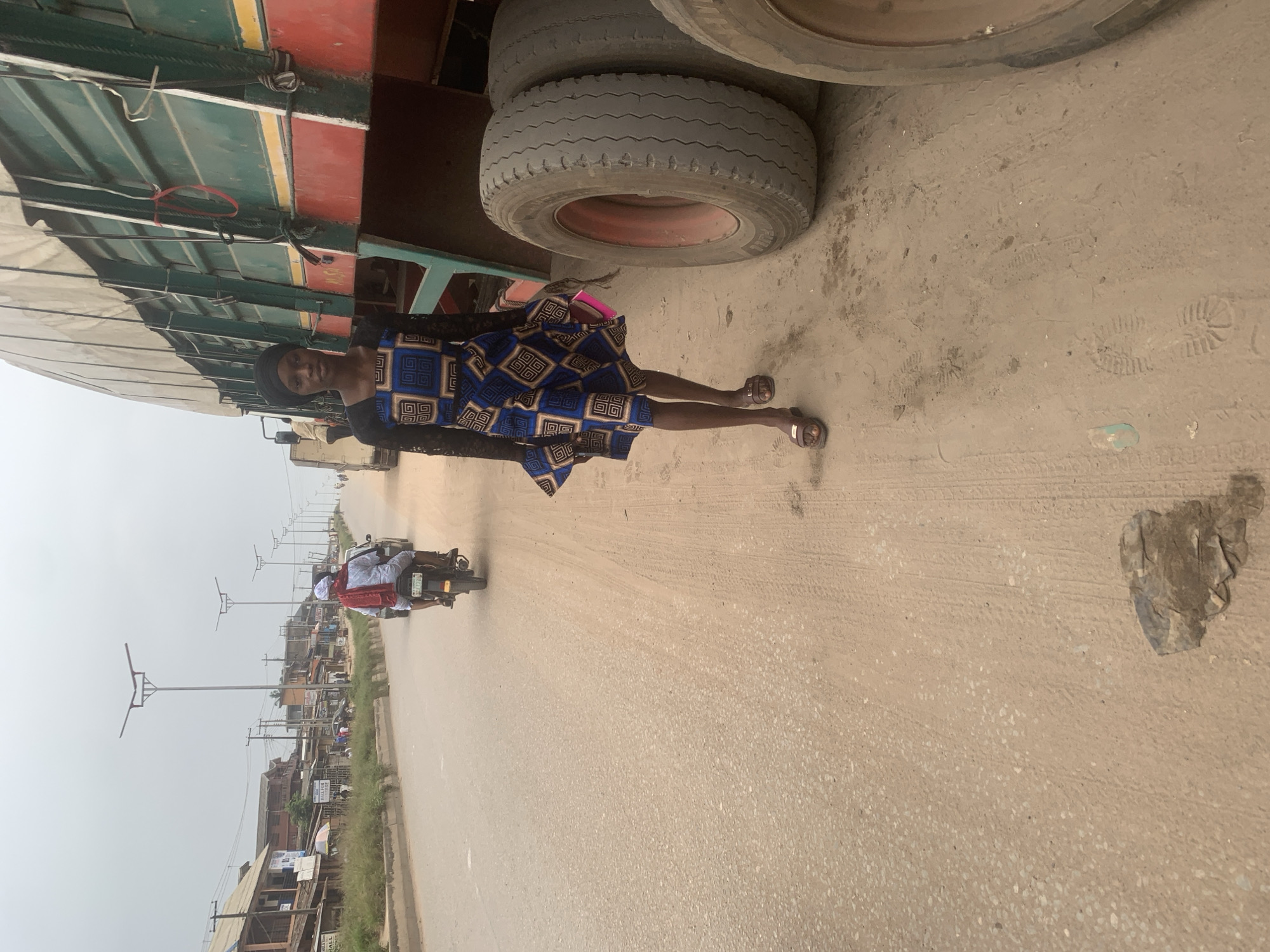 A young woman with dark skin, wearing a blue and black patterned dress, a black headscarf, and brown sandals, walks on the right side of a dusty road in Shagamu, Nigeria. She is looking towards the camera and holding a pink folder in her left hand. Beside her is a large green and red truck with its tires visible. In the background, a man wearing a white outfit and a red scarf rides a motorcycle on the left side of the road, moving away from the camera. The road is lined with buildings and streetlights. The sky is overcast.