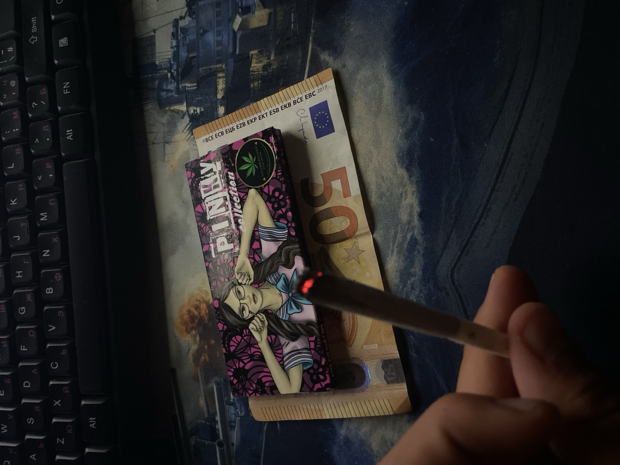 The image displays a dimly lit desk scene, observed from a first-person perspective, in the city of Unknown, Unknown. In the lower left foreground, a human hand holds a lit, hand-rolled cigarette or joint, identifiable by the glowing ember at its tip. Directly beneath this, on a desk surface, a 50 Euro banknote is visible, partially obscured by a rectangular package of rolling papers. The rolling paper package is labeled "PINKY COLLECTION" and features an illustration of a woman wearing glasses and a school uniform, along with text "KING SIZE SLIM" and a circular logo containing a cannabis leaf and the phrase "THE CANNABIS SHOP IN YOUR NEIGHBORHOOD". The surface beneath these items is a mousepad, which depicts a detailed illustration of a naval battle, showing multiple warships and plumes of smoke on a dark, turbulent sea. In the upper background, a black computer keyboard with both Latin and Cyrillic character layouts is visible, suggesting a Russian or similar Slavic language input.