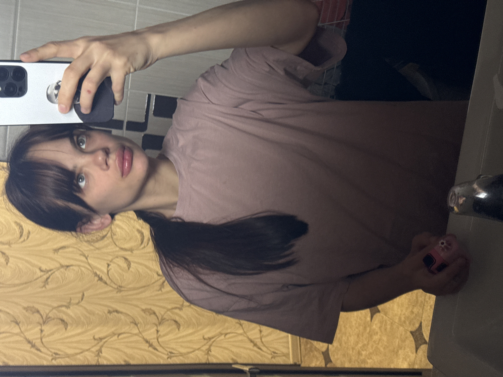 A young woman takes a selfie in what appears to be a bathroom. She is wearing a mauve t-shirt and has dark hair pulled back in a ponytail. She is holding a smartphone in her left hand with the camera pointed at her face. In her right hand, she is holding a small pink object. The background includes a tiled wall and a sink. There is no information available to confirm the location is Rostov-on-Don, Russia.