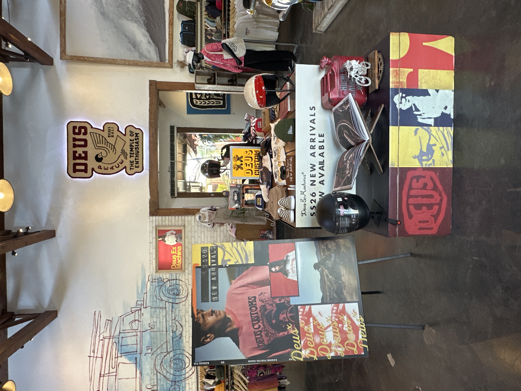 The image depicts the interior of a Deus Ex Machina store, likely in North Kuta, Indonesia, as indicated by the "80361 BALI" text on a sign. The store features displays of clothing, accessories like helmets, and promotional materials. Prominently featured are signs and displays with the "Deus Ex Machina" branding. One display advertises "SS26 NEW ARRIVALS." Artwork and graphics adorn the walls, including a large drawing of a vehicle and other various pictures.