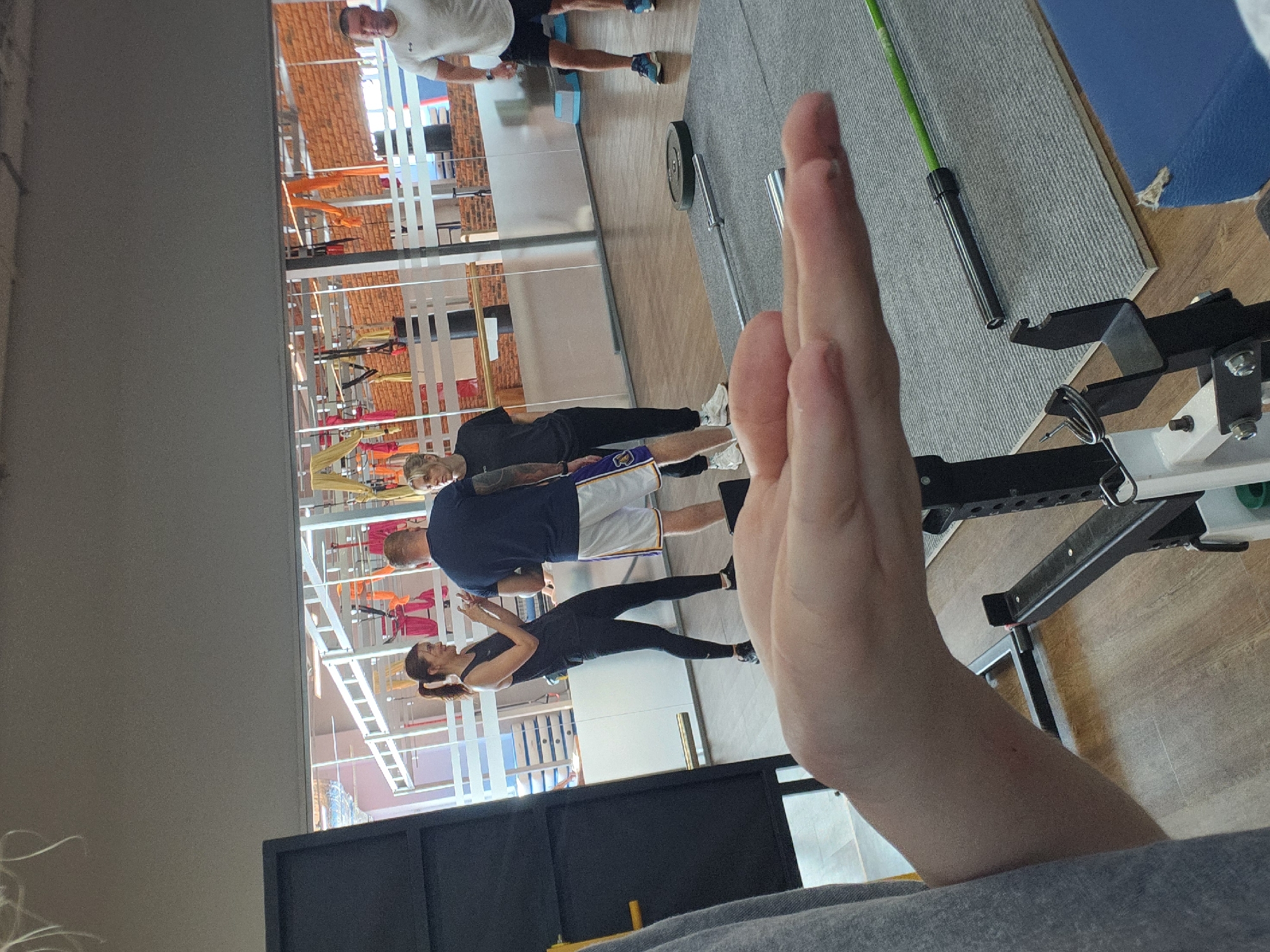 The image shows an indoor gym setting with exercise equipment and people visible through a glass wall. The gym features exposed brick walls, a white ceiling with suspension equipment, and various fitness training areas. Four individuals are observable: one man in white athletic wear, one man in a navy shirt and white shorts, a woman in black athletic wear, and another man in a black sweatshirt and purple and gold shorts. The hand of the photographer is prominently featured in the foreground. A barbell and bench press setup can be seen in the nearby space. The location is in the city of Unknown, Unknown.