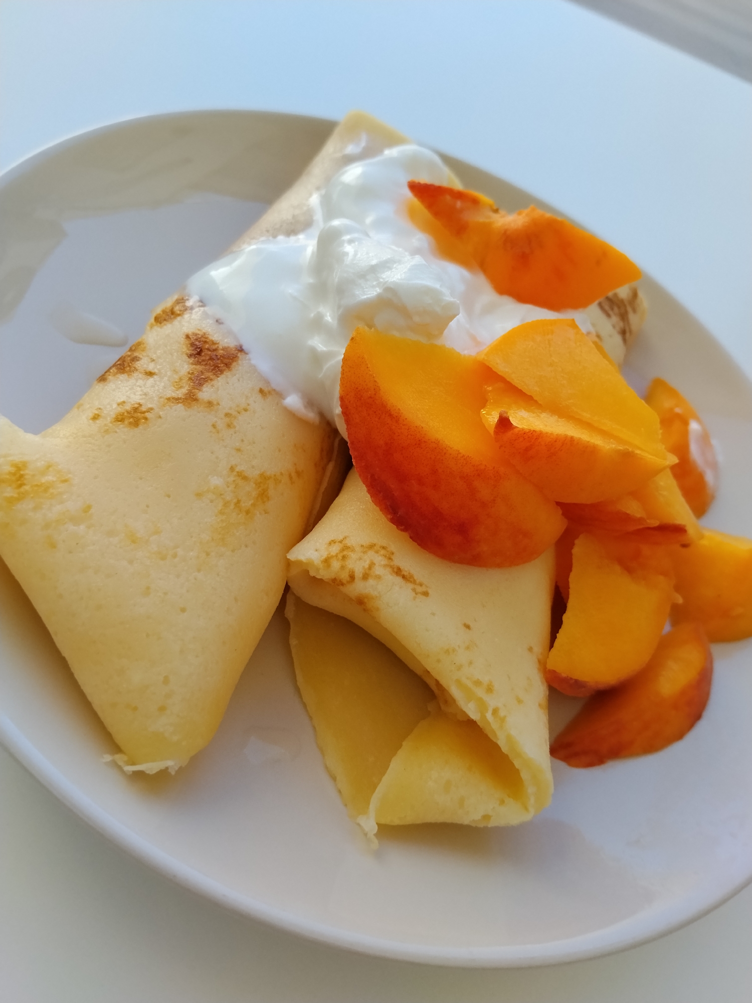 A close-up, high-angle shot shows a plate of crepes topped with whipped cream and sliced peaches. The crepes are golden brown and folded, with visible specks of browning. A dollop of fluffy white whipped cream sits atop the crepes, and several bright orange peach slices are arranged around them. The food is presented on a white plate, which is placed on a white surface that appears to be a table. The lighting suggests an indoor setting with natural light. The scene depicts a meal, likely breakfast or dessert, in Bitonto, Italy.