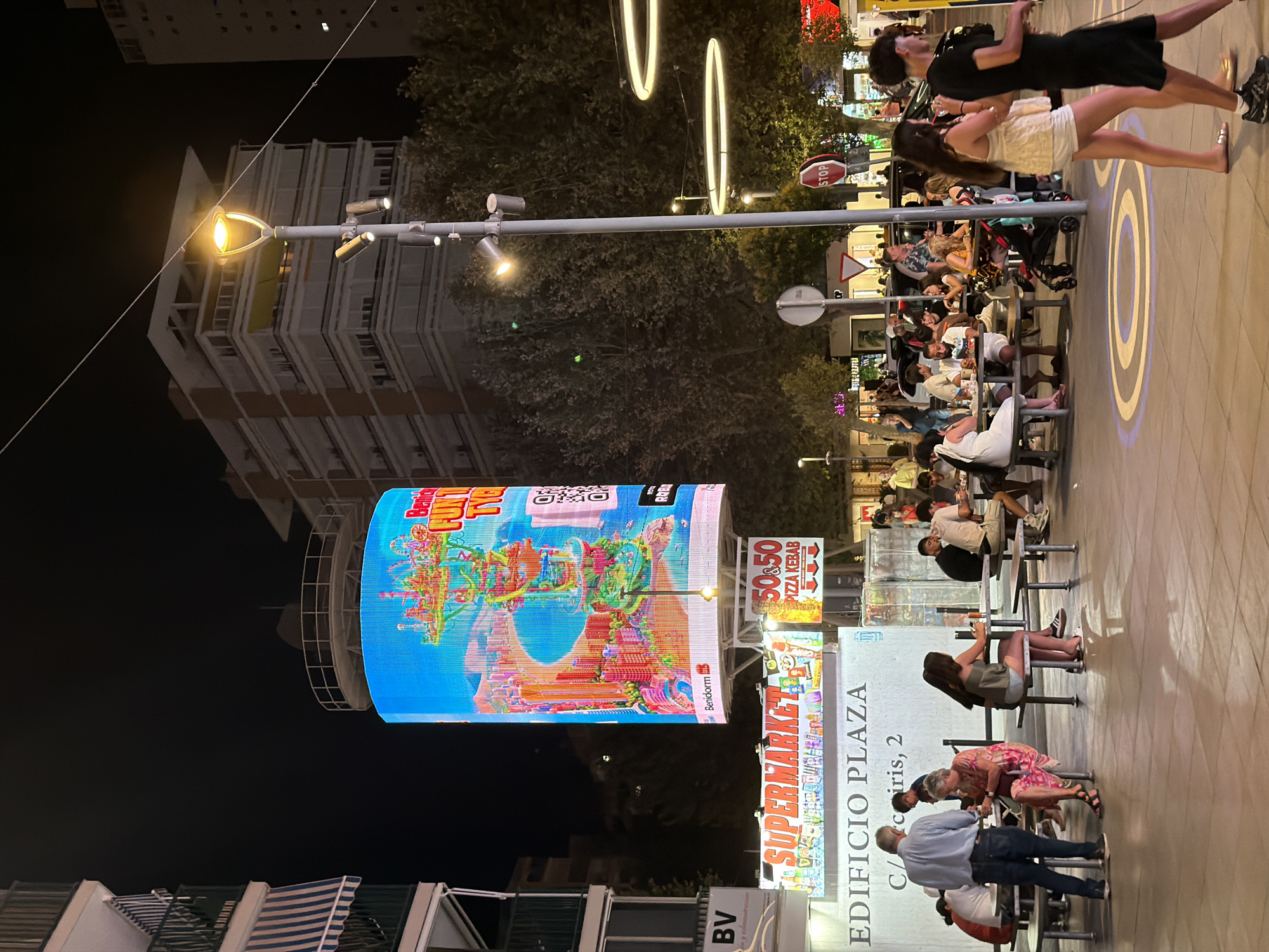 The image shows a nighttime scene in Benidorm, Spain, likely in a busy tourist area with outdoor seating. A large, illuminated digital billboard displays a vibrant, colorful image of what appears to be a theme park or attraction with a beach and cityscape. Below the billboard, a sign reads "SUPERMARKET," and another smaller sign advertises "50 & 50 PIZZA KEBAB." An area labeled "EDIFICIO PLAZA C/ de Ciris, 2" suggests the location. Several people are seated at tables under the sign, engaged in dining or conversation. Others are walking through the area, including a couple in the foreground. The environment is urban, with trees, streetlights, and buildings visible. The overall atmosphere is lively and commercial.