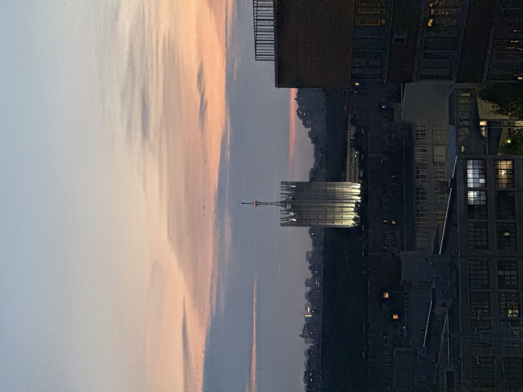 A wide-angle, eye-level shot captures an urban cityscape at dusk. The sky transitions from a pale blue at the top to soft hues of pink and orange near the horizon, with wispy clouds streaking across. Dominating the middle ground is a tall, art deco-style tower, illuminated from below, its tiered structure topped with a radio antenna. In the foreground and extending into the middle ground, various multi-story buildings are visible, some with lights glowing from their windows. A large, dark building with a balcony railing is prominent on the right side of the frame. The overall impression is of a city settling into evening, with the last light of day illuminating the sky above the urban sprawl. The specific location in the city of Unknown, Russia, is not identifiable from the image itself.