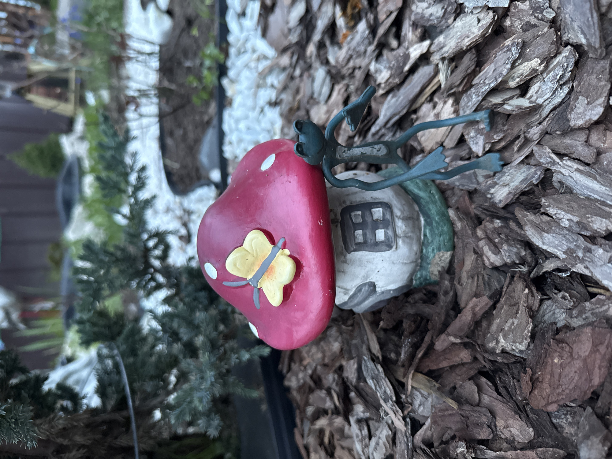 The image shows a garden decoration consisting of a mushroom-shaped house with a red roof and a yellow butterfly on top, placed on a bed of bark mulch. The house is white with a small window. The scene appears to be outdoors. The city and country are irrelevant as this is a photograph of an object not location.