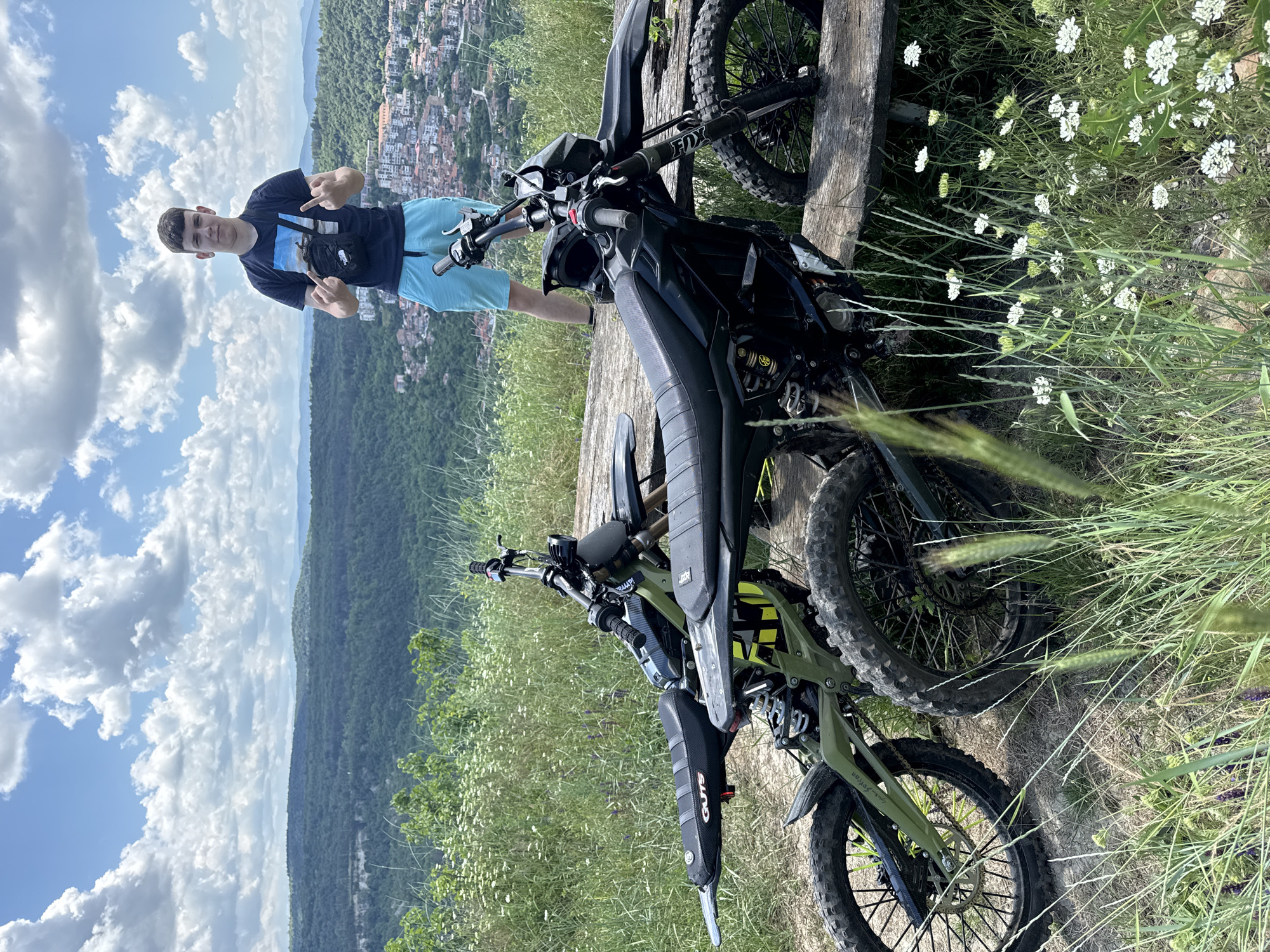 A young man stands in front of two electric dirt bikes, outdoors with a panoramic view of the city of Veliko Tarnovo, Bulgaria, in the background. He is wearing a dark blue t-shirt, light blue shorts, and a black shoulder bag. He is giving the middle finger to the camera with both hands. The dirt bikes are parked on a wooden surface that appears to be a bench or platform. The environment is grassy with wildflowers and green trees. The sky is cloudy and blue.