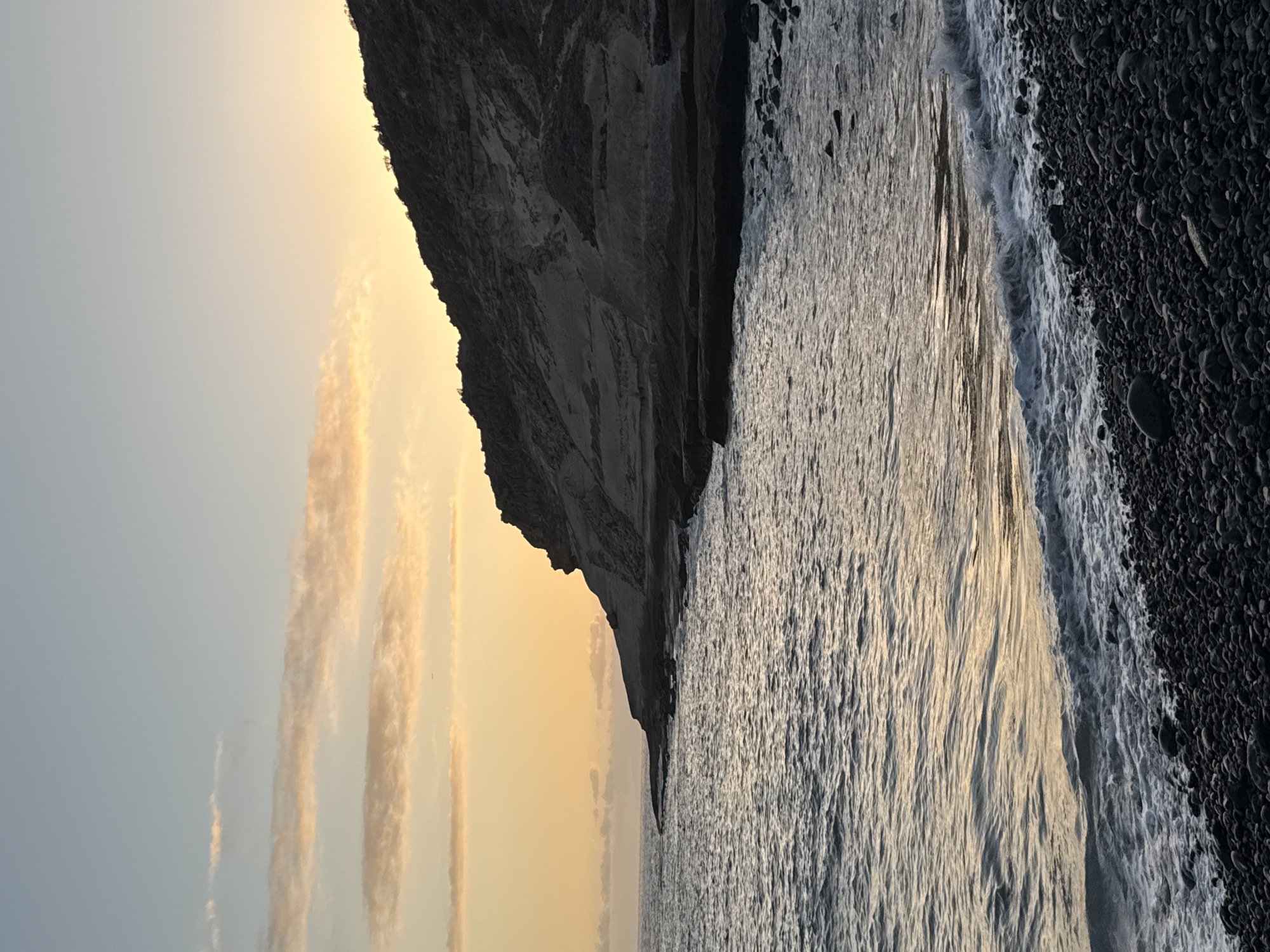 The image depicts a rocky coastline and the ocean at sunset in Adeje, Spain. A dark, imposing cliff face dominates the right side of the frame, with its jagged contours softened by the twilight. The sea is calm, with gentle waves lapping against the shore, which is composed of dark pebbles. The sky above transitions from a pale blue at the top to a warm orange hue near the horizon, streaked with wispy, pink-tinged clouds. The overall scene evokes a sense of natural beauty and tranquility.