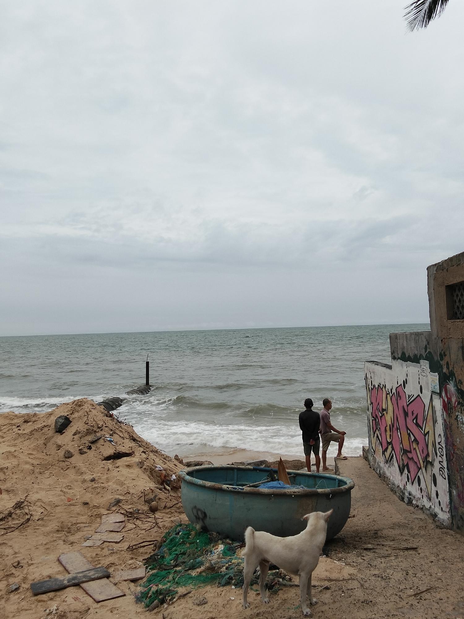 The image depicts a cloudy day at a beach in Phan Thiết, Vietnam, with the ocean in the background. In the foreground, a white dog stands on a sandy beach, facing away from the camera. To the left, a large pile of sand contains debris. A blue, boat-shaped object is present on the sand near the dog. In the middle ground, two men, one wearing a black shirt and shorts, and the other wearing a light-colored polo shirt and shorts, stand near the water's edge, facing the ocean. To the right, a concrete structure with graffiti on its wall is visible. The ocean waves are visible lapping at the shore. A dark, vertical post in the water is seen in the distance.
