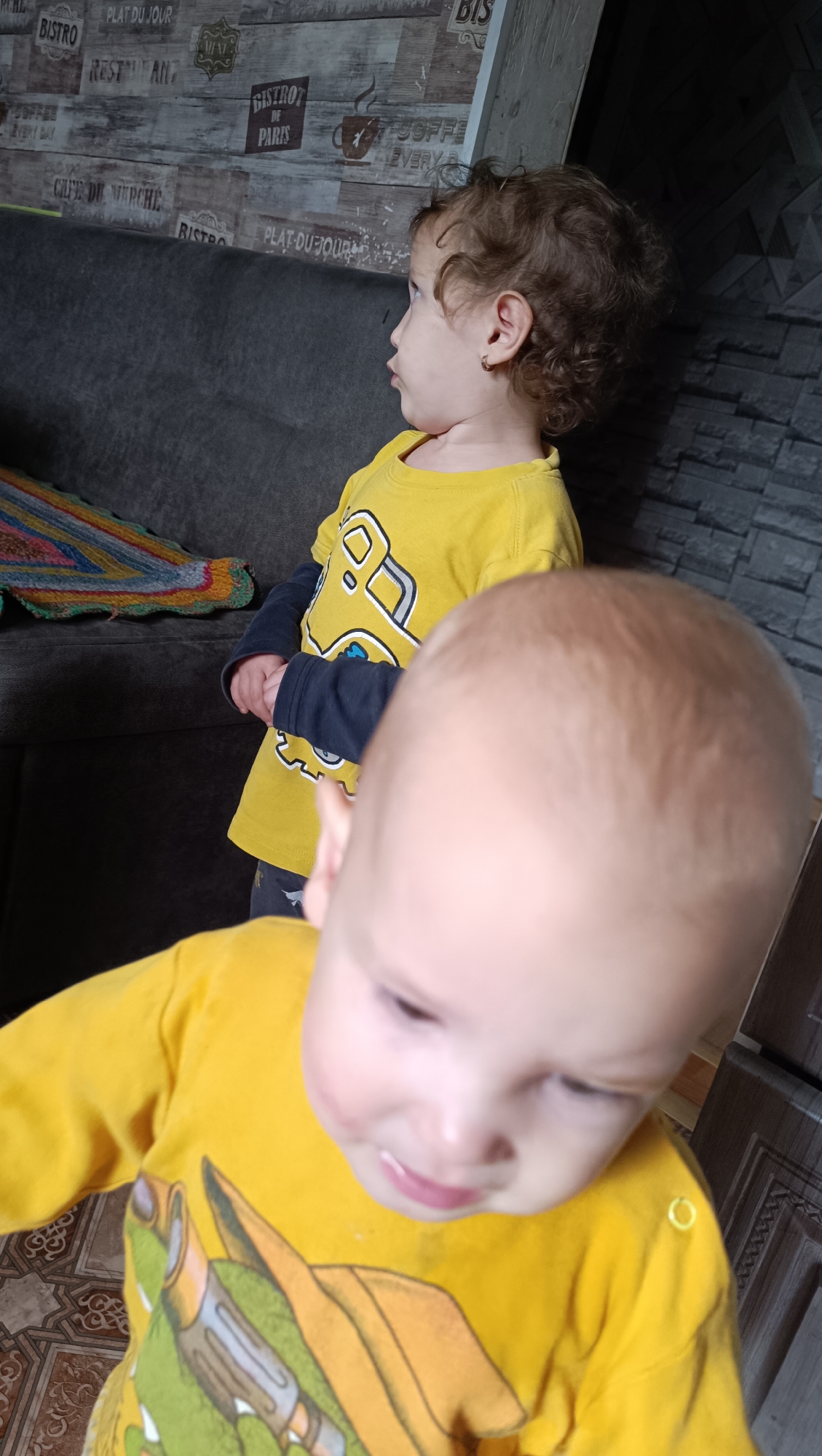 The image shows two young children indoors, one in the foreground and one in the background. The child in the foreground is facing the camera, with their head and upper body visible. They are wearing a yellow long-sleeved shirt with a graphic of a dragon. The child in the background is facing away from the camera, in profile, looking to the left. They have curly brown hair and are wearing a yellow long-sleeved shirt with a graphic of a truck. The background includes a dark sofa with a colorful knitted item on it, a wall with a wood-grain pattern and some text, and a textured gray wall. The floor appears to be tiled. The lighting suggests an indoor setting, possibly a home. The provided location information "city of Karsha, Russia" is not supported by any visual evidence in the image itself.