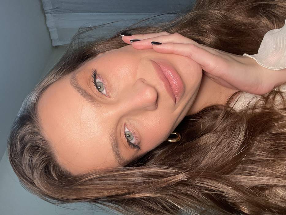 A woman with long brown hair poses, holding her hand near her face. Her eyes are green. She has on eyeliner and lip gloss, and her nails are painted black with silver tips. She's wearing a gold hoop earring and a white ruffled top. The background is white and indistinct.